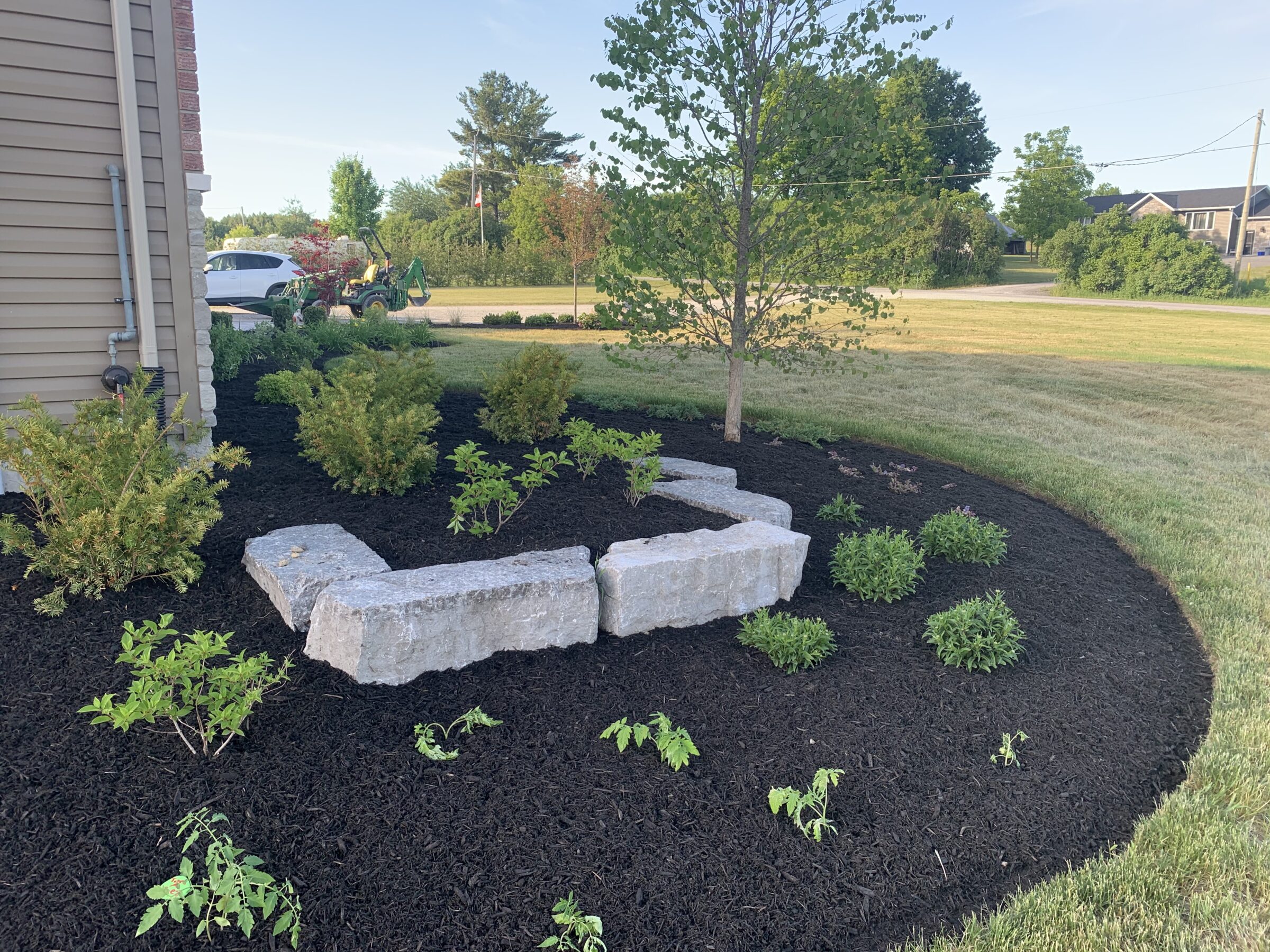 A landscaped garden with mulch and stone features beside a building, near grass, trees, a car, and a green utility vehicle.