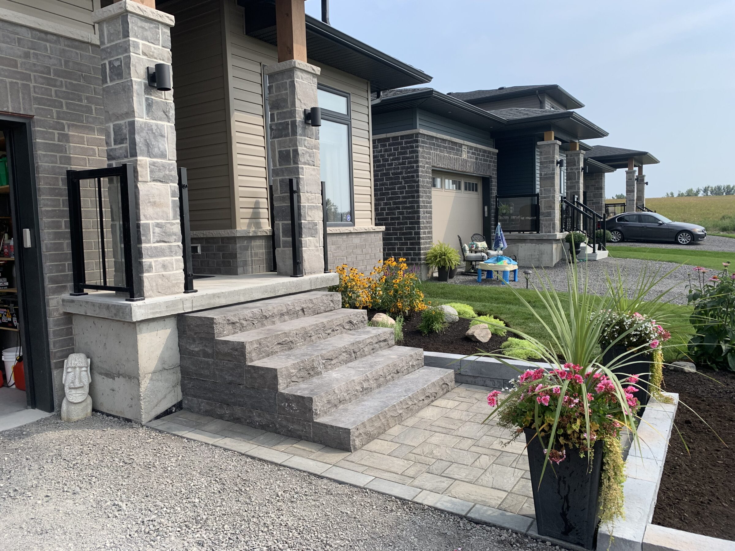 Modern brick house with landscaped garden, gray car on driveway, and stone steps leading to porch. Potted plants and decorative rocks enhance curb appeal.