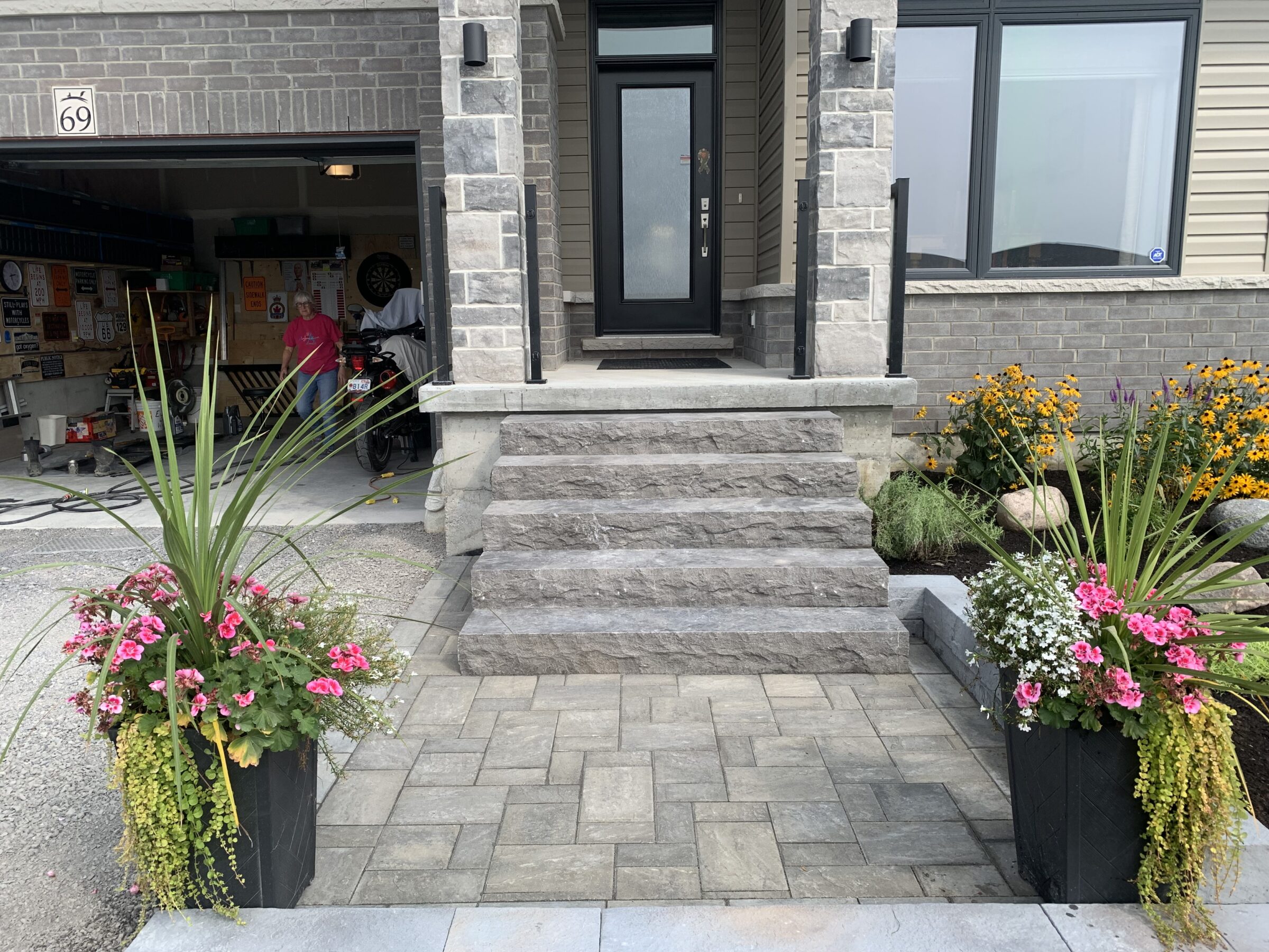 A house entrance with stone steps, flanked by potted flowers, opens to a garage where a person and motorcycles are visible.