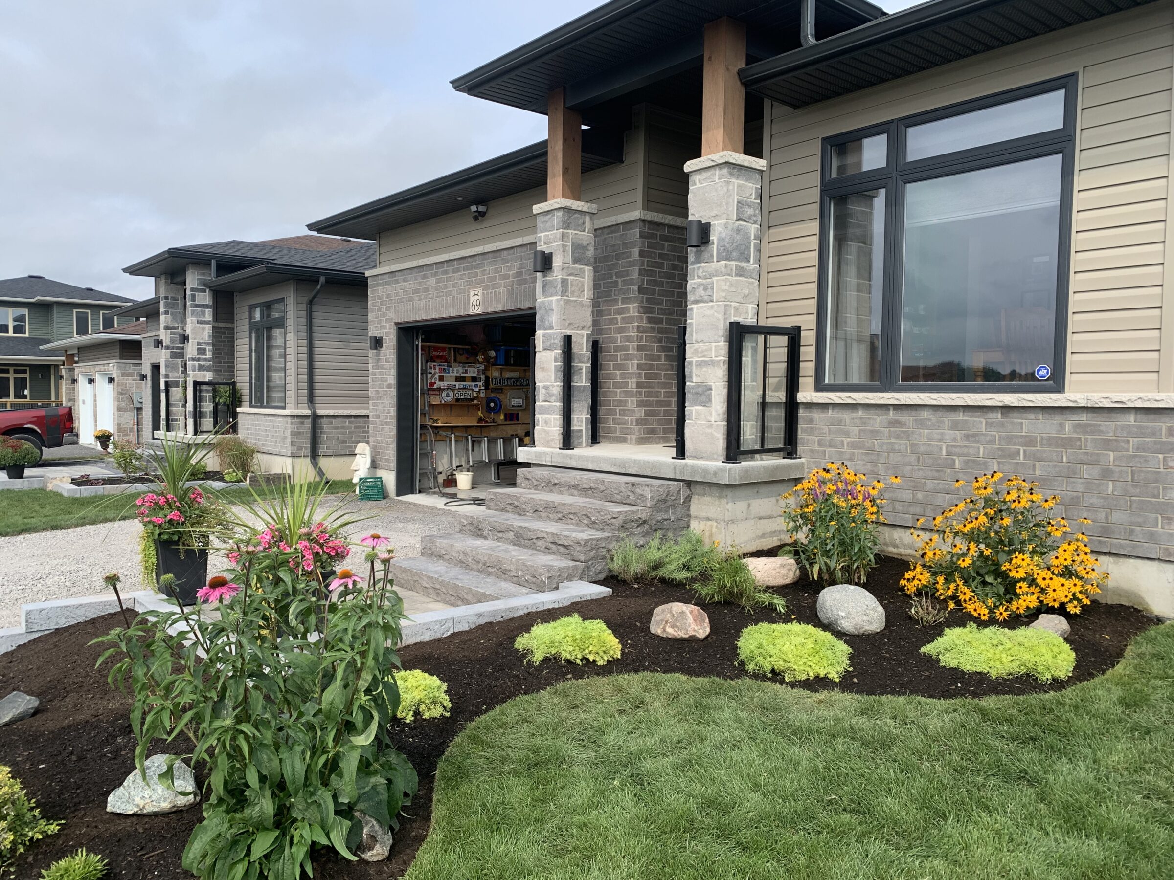 Modern house exterior with a manicured garden, open garage displaying tools, and neatly trimmed lawn, set in a suburban neighborhood.