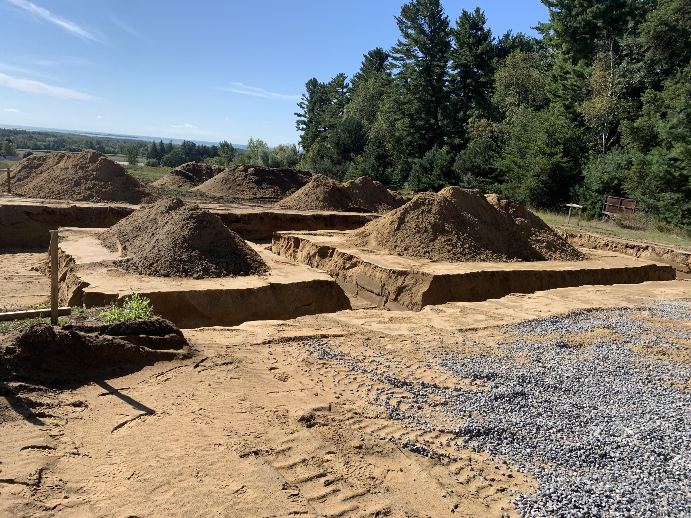 A construction site with large sand piles surrounded by dense green trees under a clear blue sky, no landmarks visible.