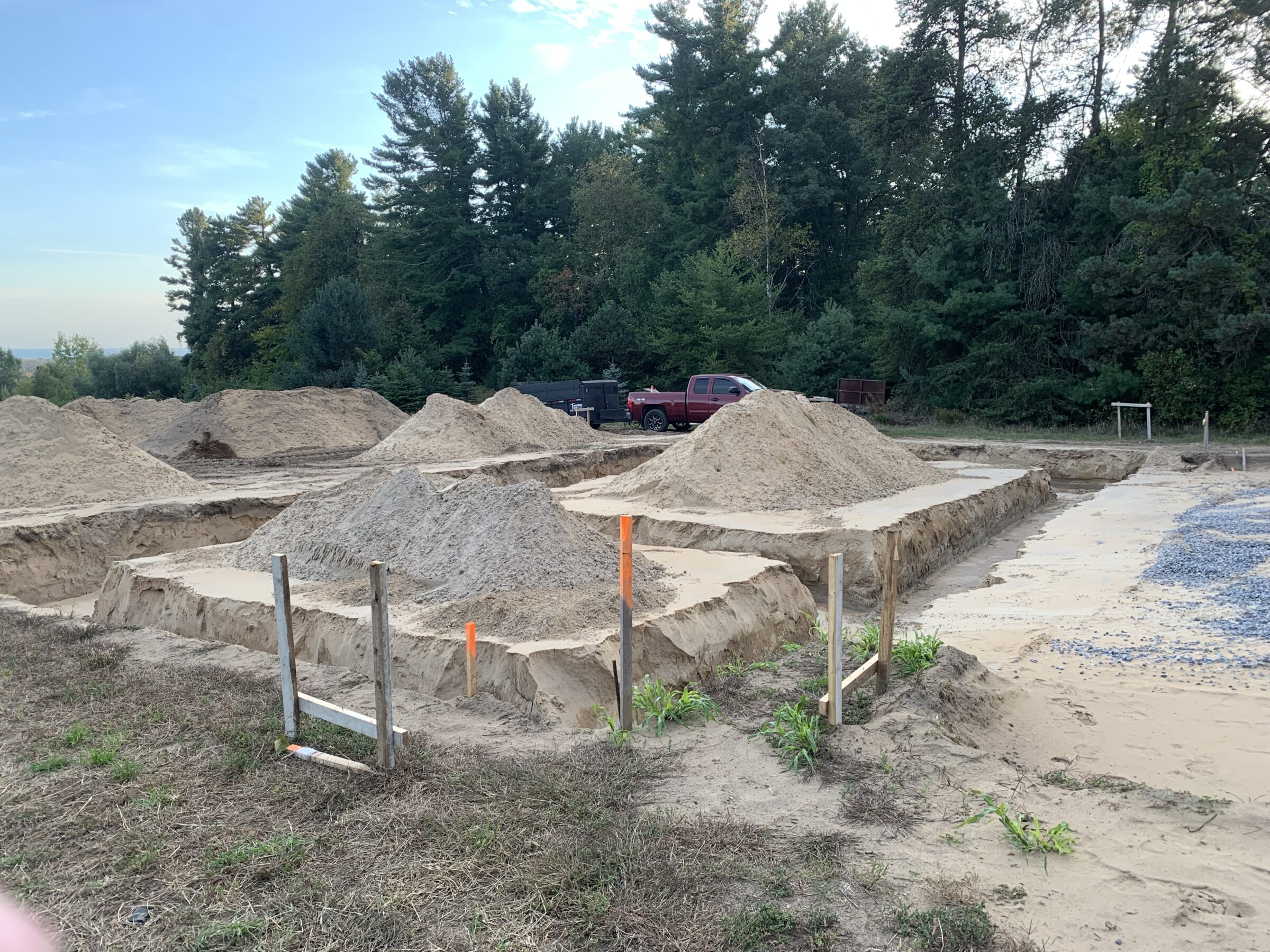 A construction site with mounds of sand, surrounded by trees. A red truck is parked in the background under a clear sky.