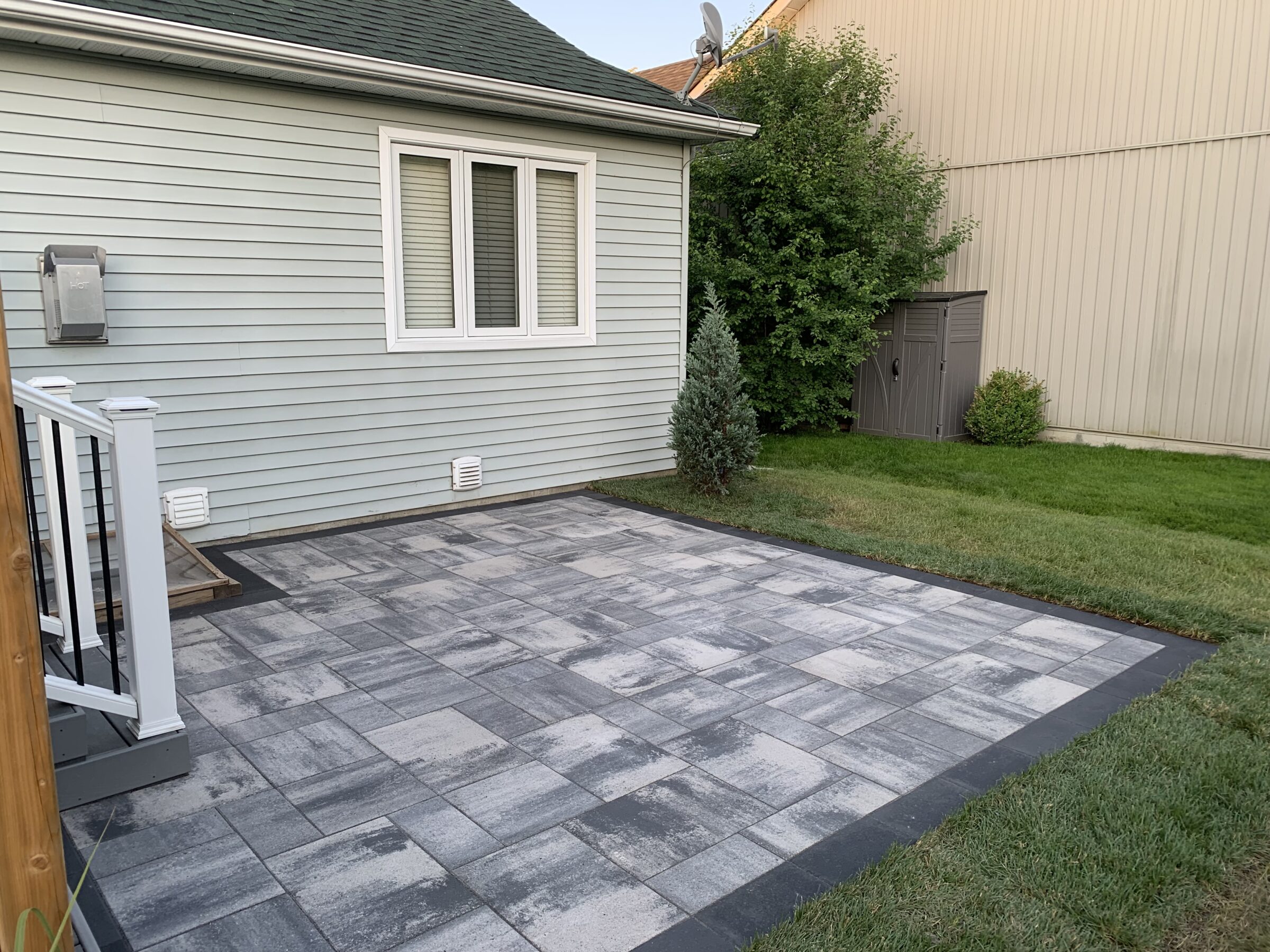 A backyard with a gray tile patio, green grass, small trees, a small storage shed, and a light blue siding building.