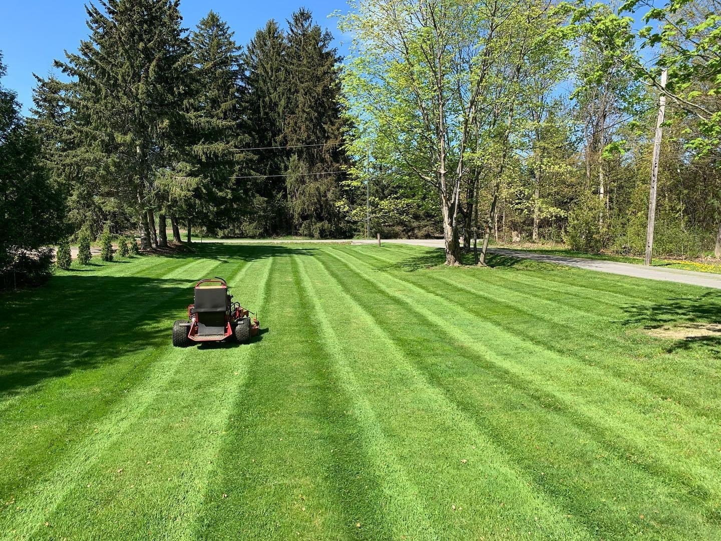 A lawn mower creating neat stripes on a freshly cut grass yard, surrounded by trees and a clear blue sky.