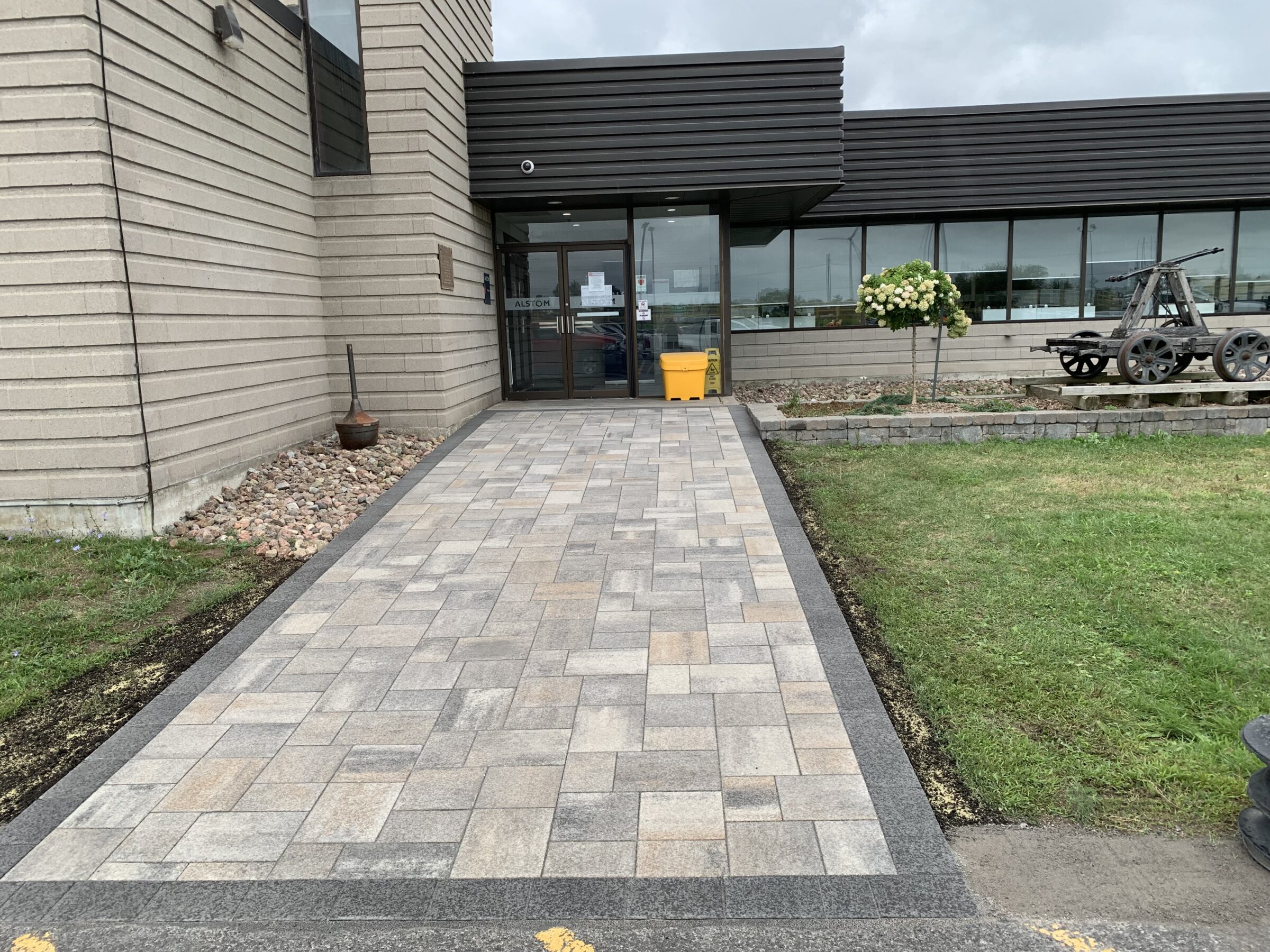 A modern building entrance with a paved walkway, landscaped greenery, and an old wooden cart displayed beside the pathway, under a cloudy sky.