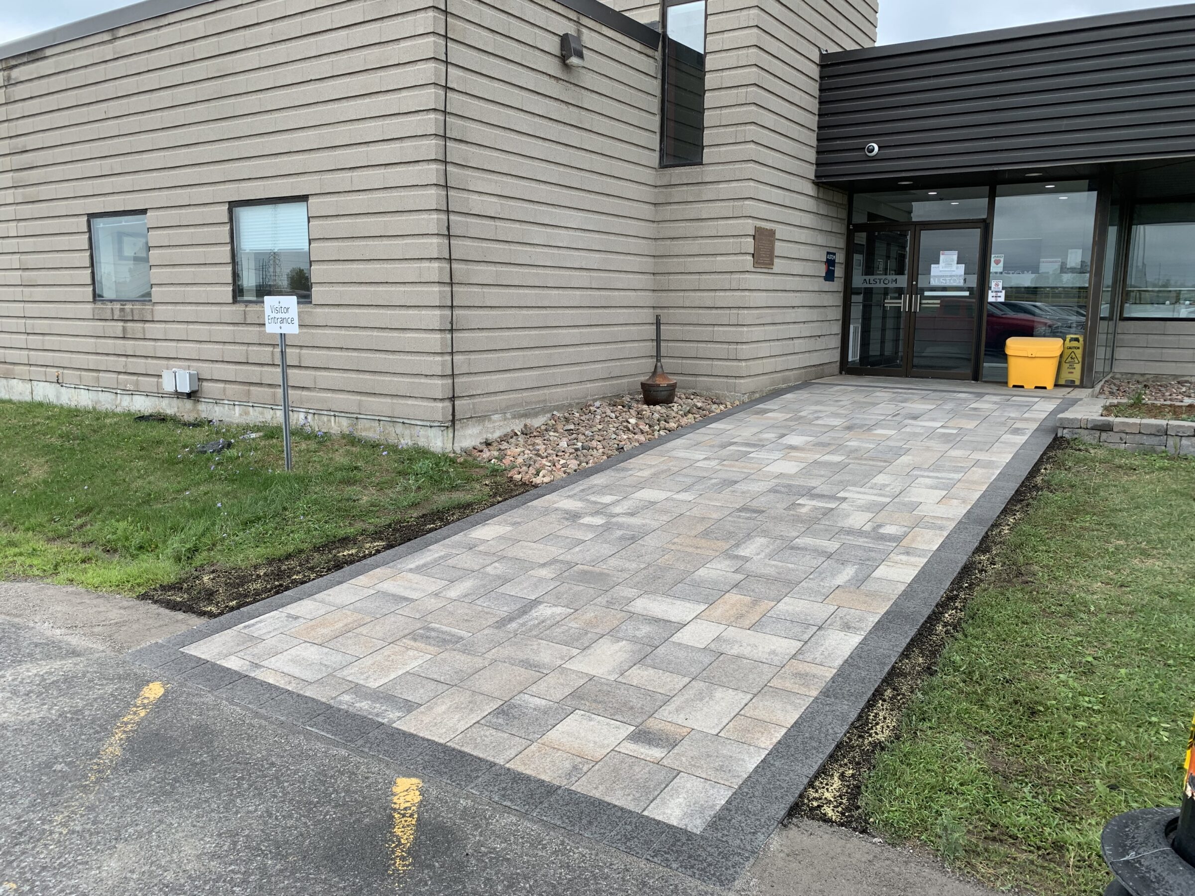 A modern building entrance with a tiled walkway, sign reading "Visitor Entrance," grass, and potted plants; glass doors and a yellow bin visible.
