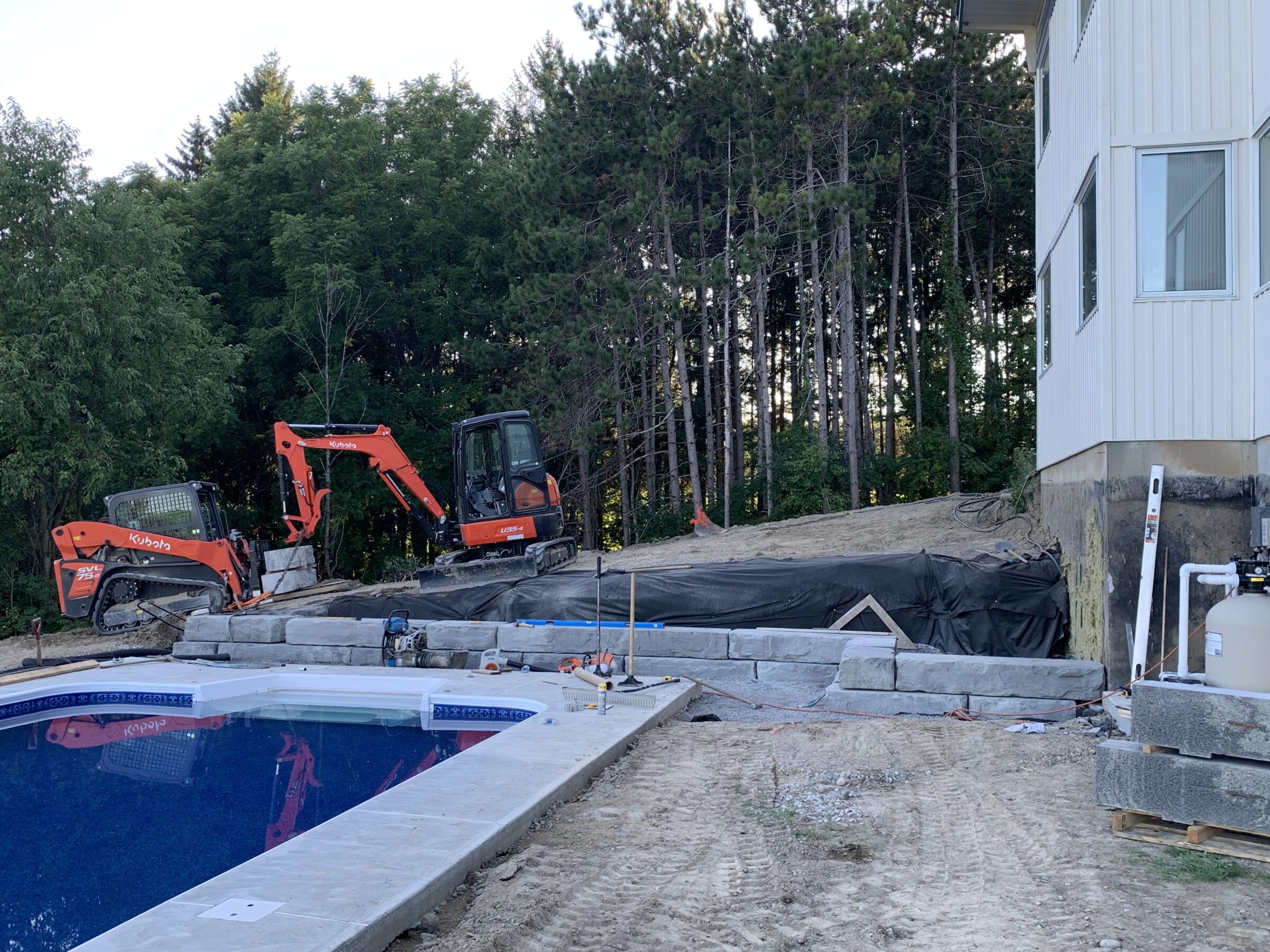 Construction site beside pool, featuring two orange excavators on a dirt hill. Surrounded by trees and a partially built retaining wall.