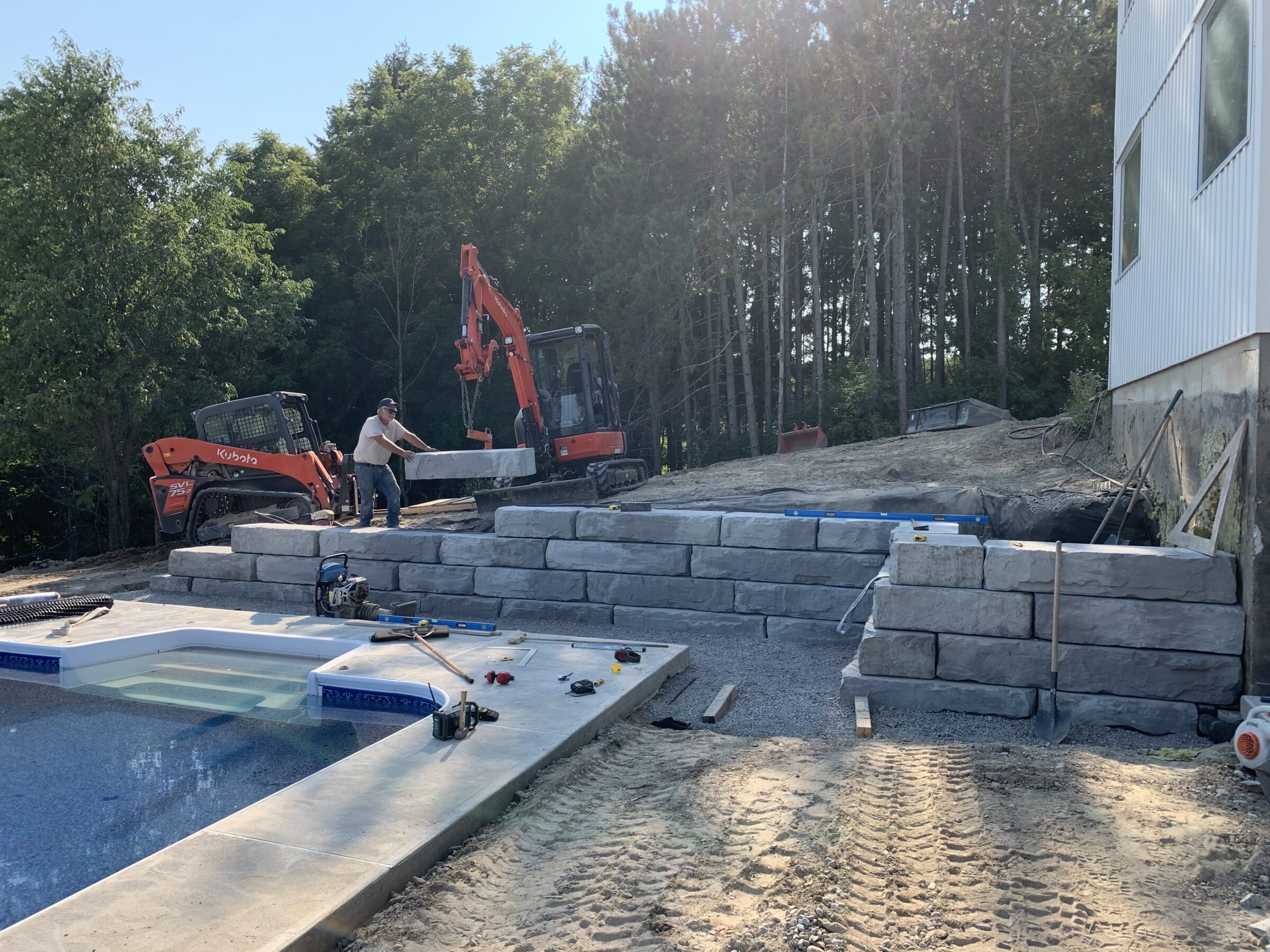 A person uses construction equipment to build a stone retaining wall near a swimming pool, surrounded by trees and a building.