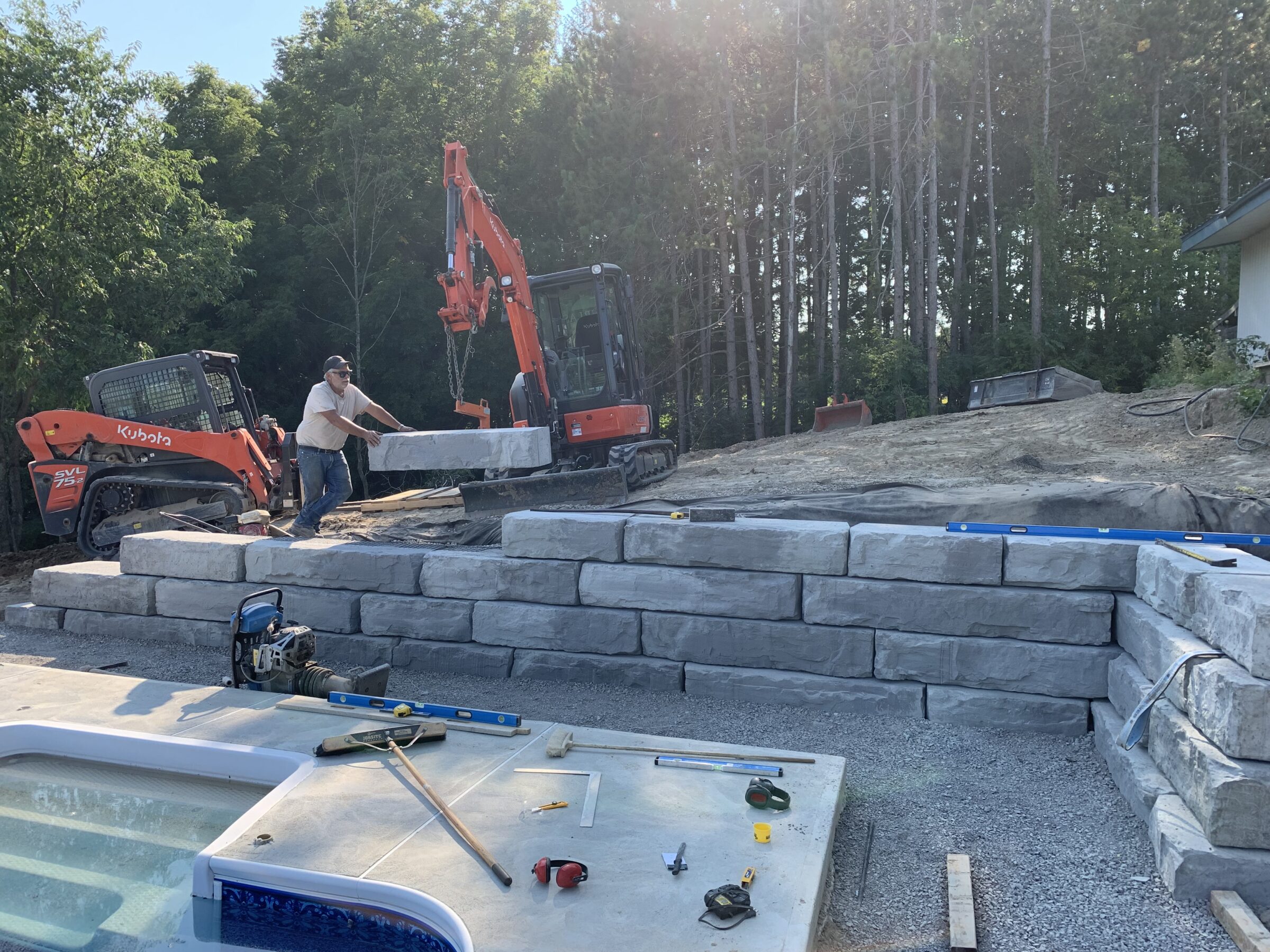 A person operates machinery, constructing a stone retaining wall near a pool, surrounded by trees and construction tools.
