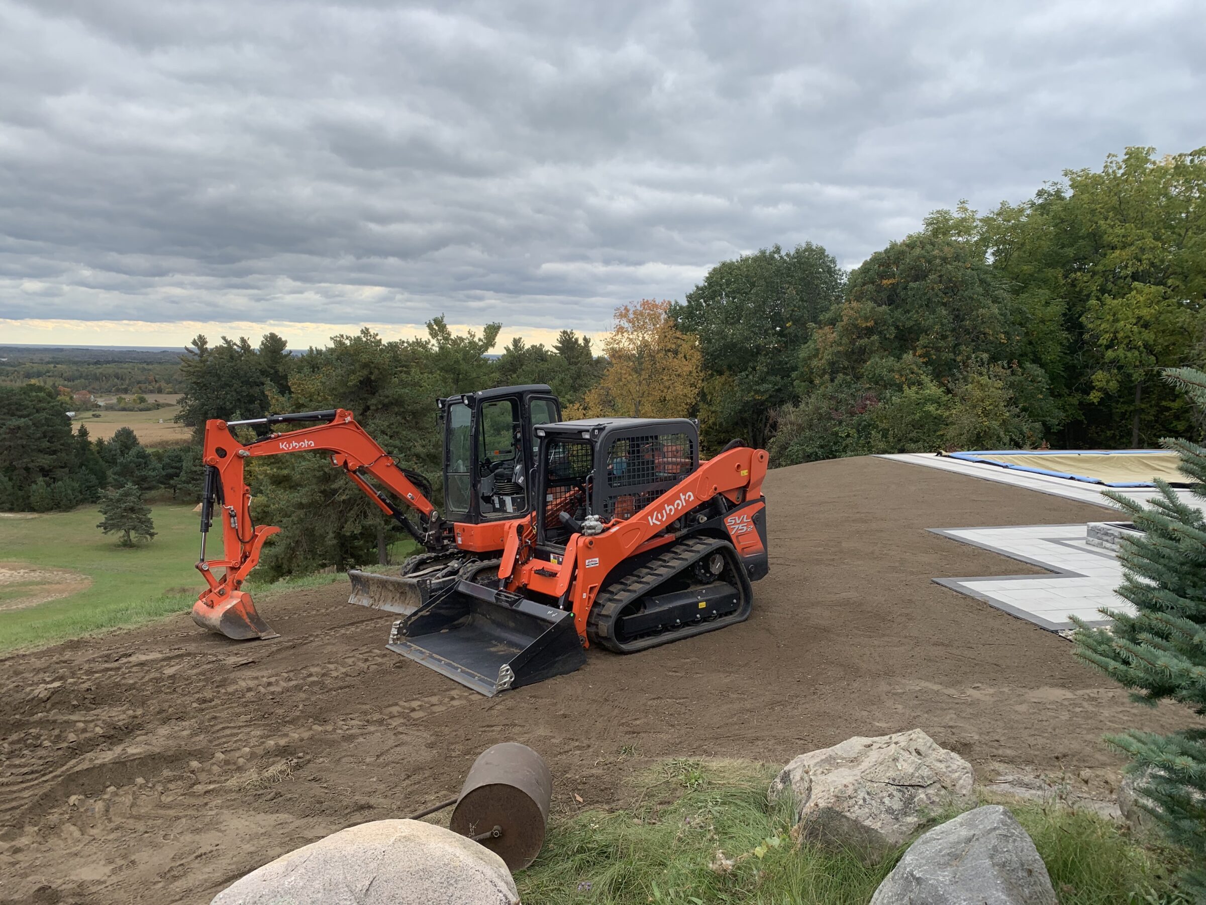 An orange excavator operates on a dirt surface surrounded by trees under a cloudy sky, with no recognizable landmarks or buildings in view.