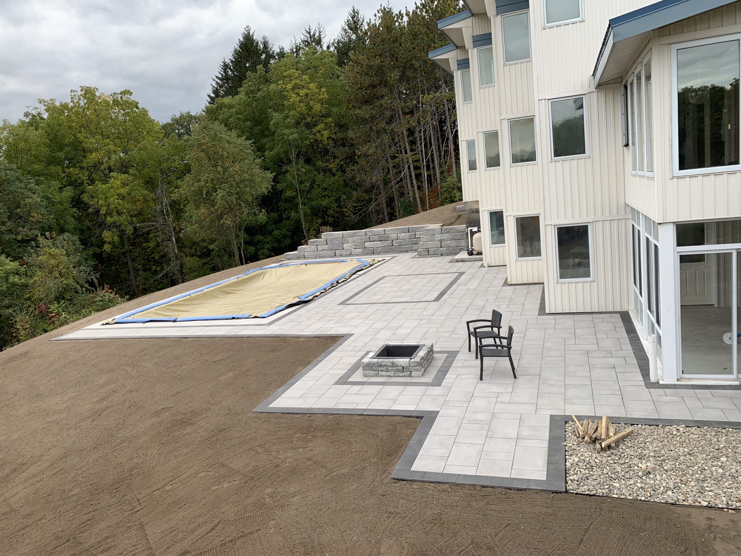 Modern house with large windows overlooks partially tiled patio, small fire pit, and covered pool, surrounded by dense green forest. Overcast sky above.