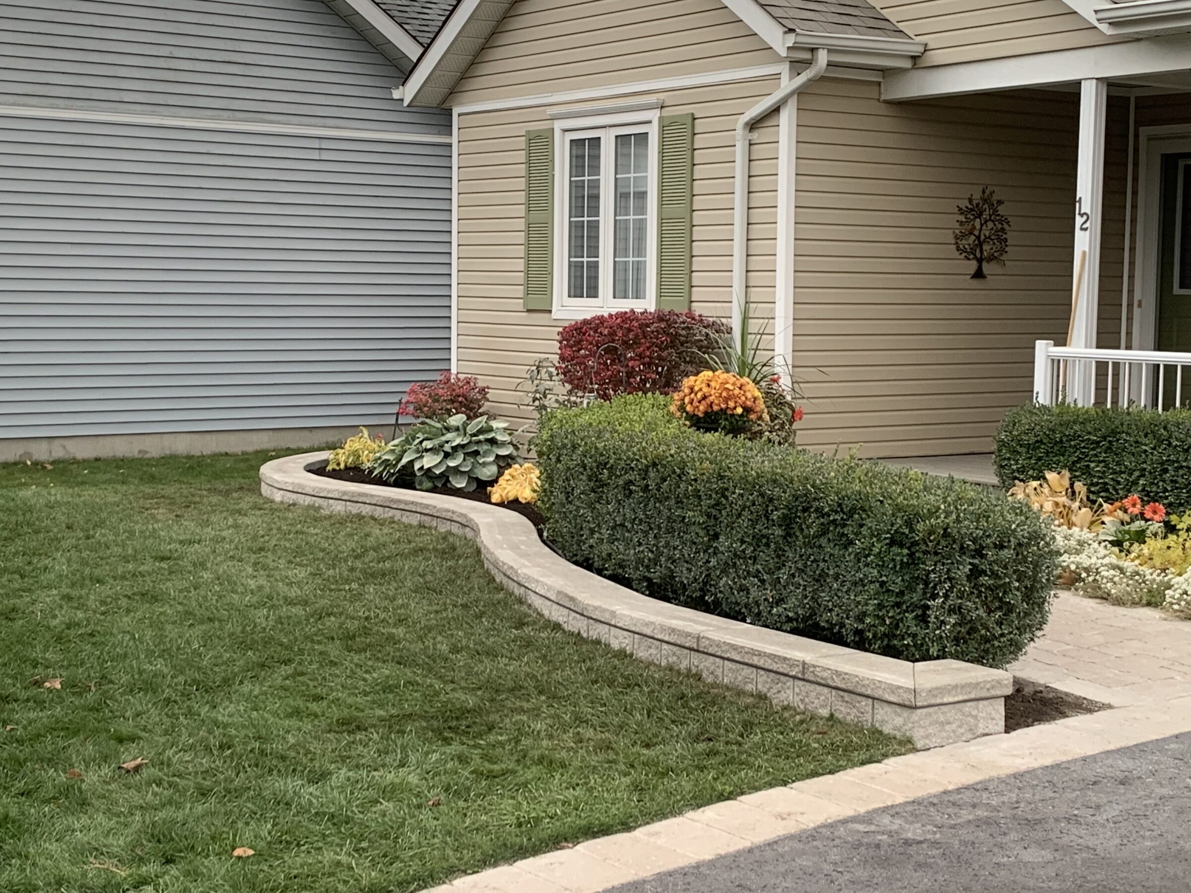 A well-maintained garden features colorful bushes and flowers in front of a beige house with green shutters and a small porch.