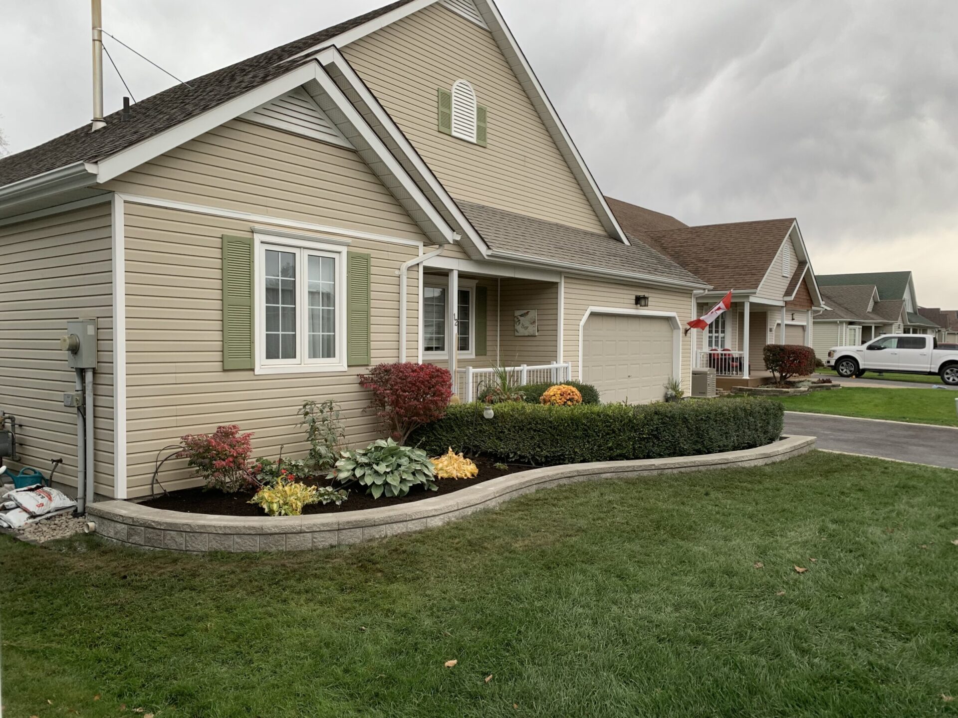 A beige suburban house features a Canadian flag, manicured garden, and quiet driveway with a white truck parked, under a cloudy sky.