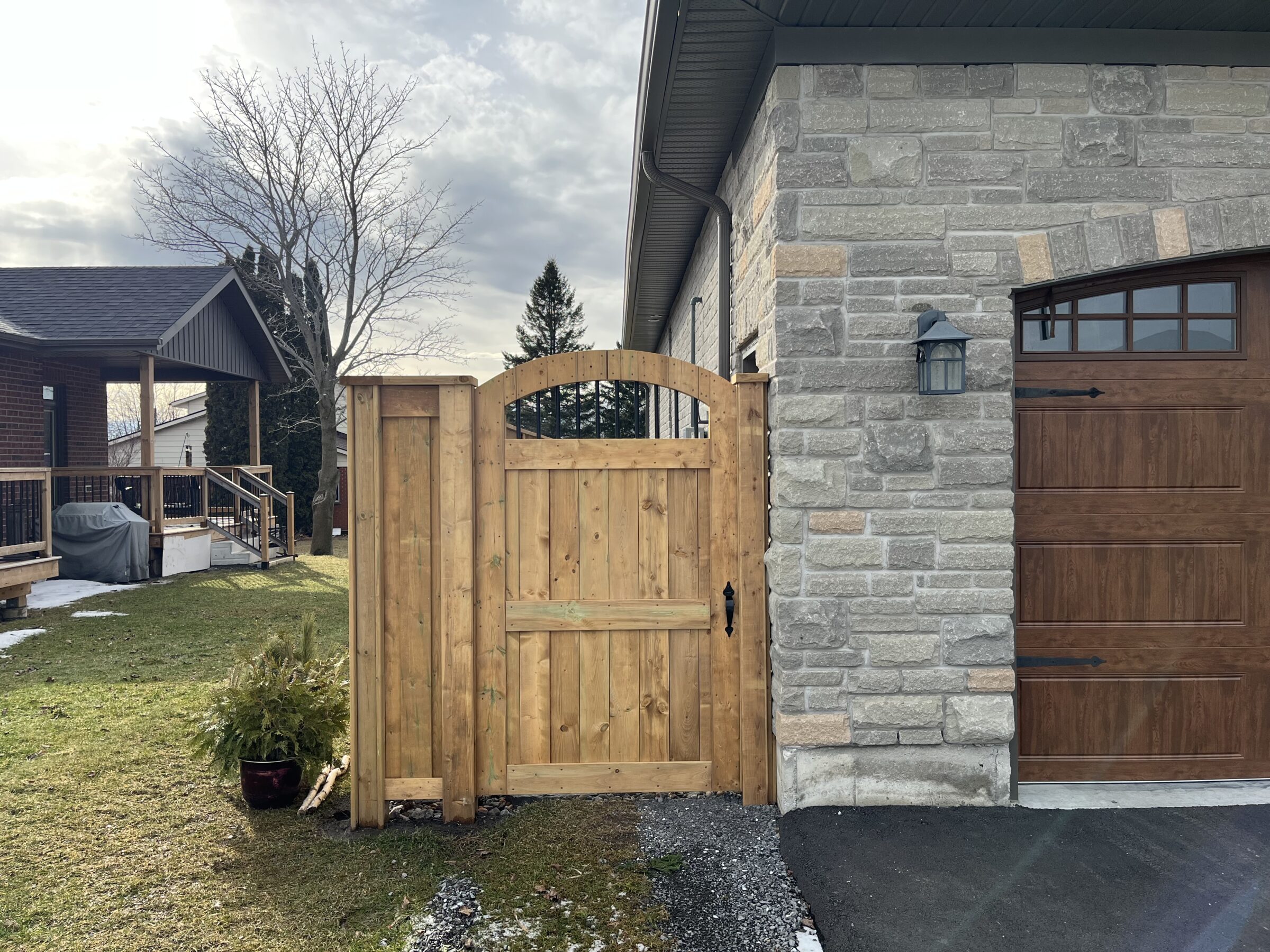 Stone house with wooden gate and garage door, adjacent to a bare tree and neighboring porch, under a cloudy sky. No recognizable landmarks.