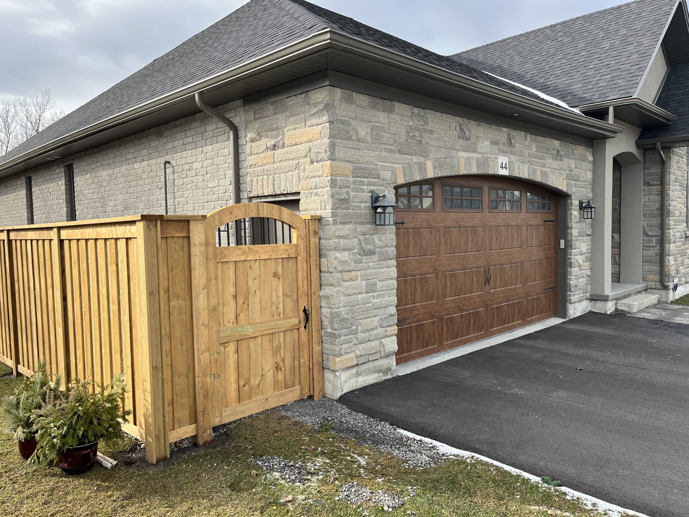 A suburban stone house features a wooden garage door, lantern-style lights, and a wooden fence with a matching gate, set in a neat lawn.