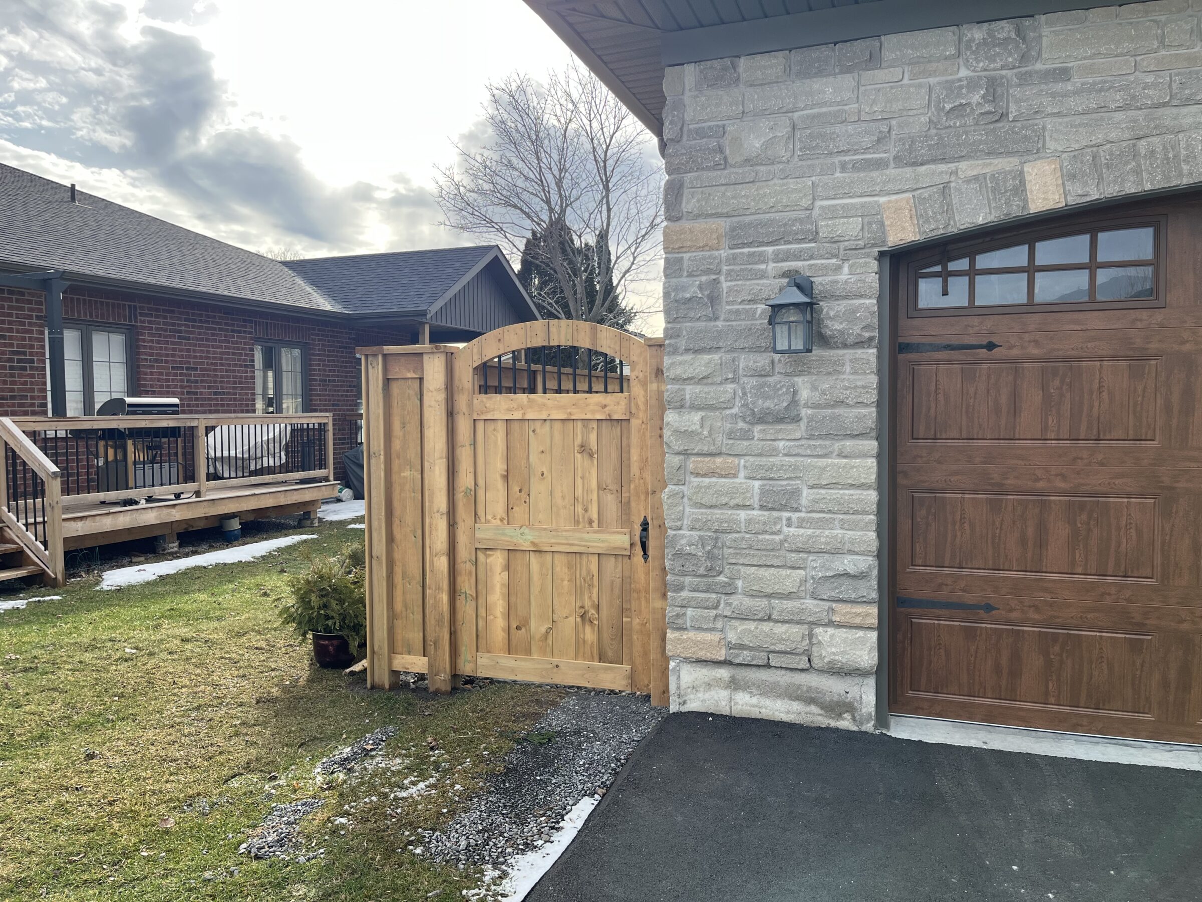 A backyard area features a wooden deck and gate, adjacent to a stone and wood garage, under a cloudy sky.