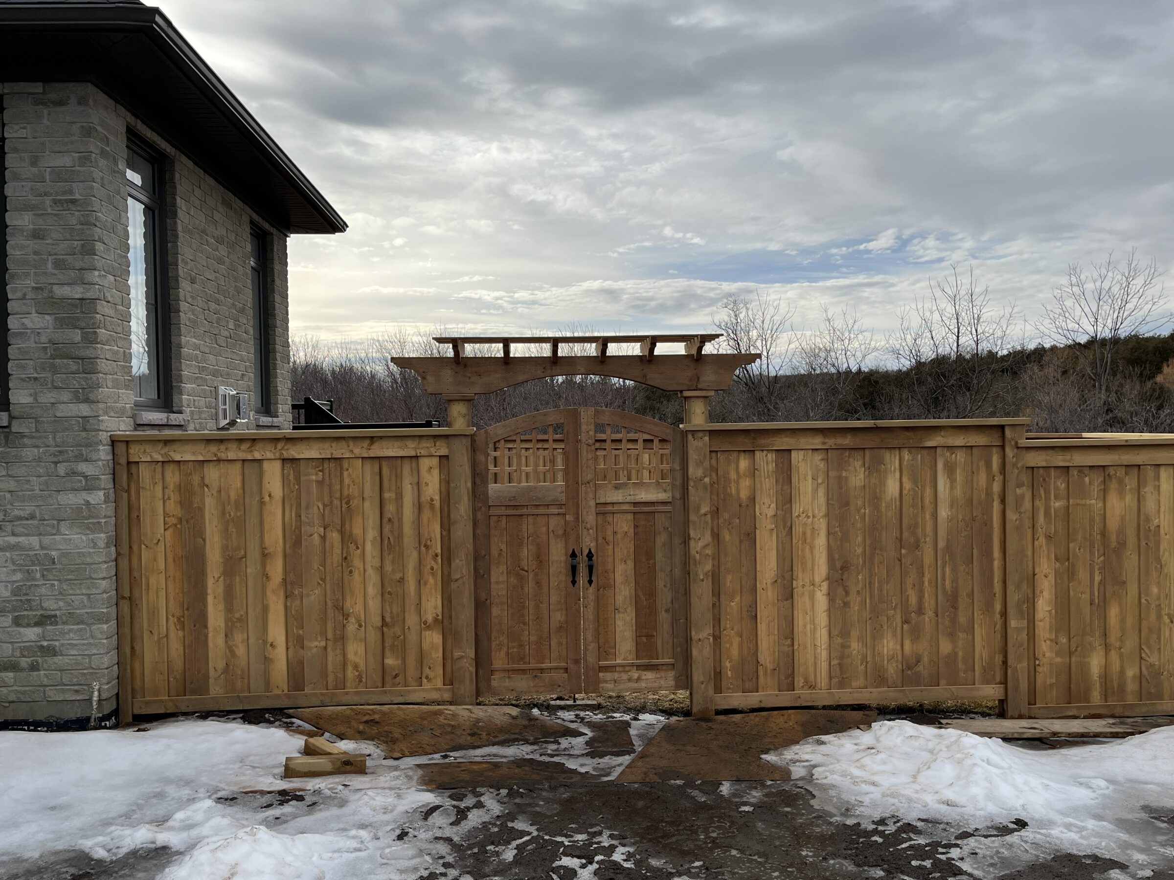 Wooden fence with gate beside a brick building. Snow partially covers the ground on a cloudy day, with bare trees in the background.