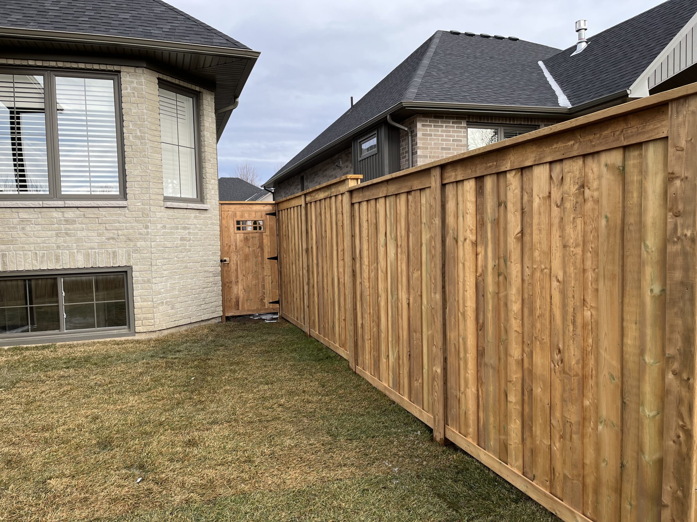 Two houses with beige brick walls are separated by a wooden fence and gate, visible on a grassy backyard under a cloudy sky.