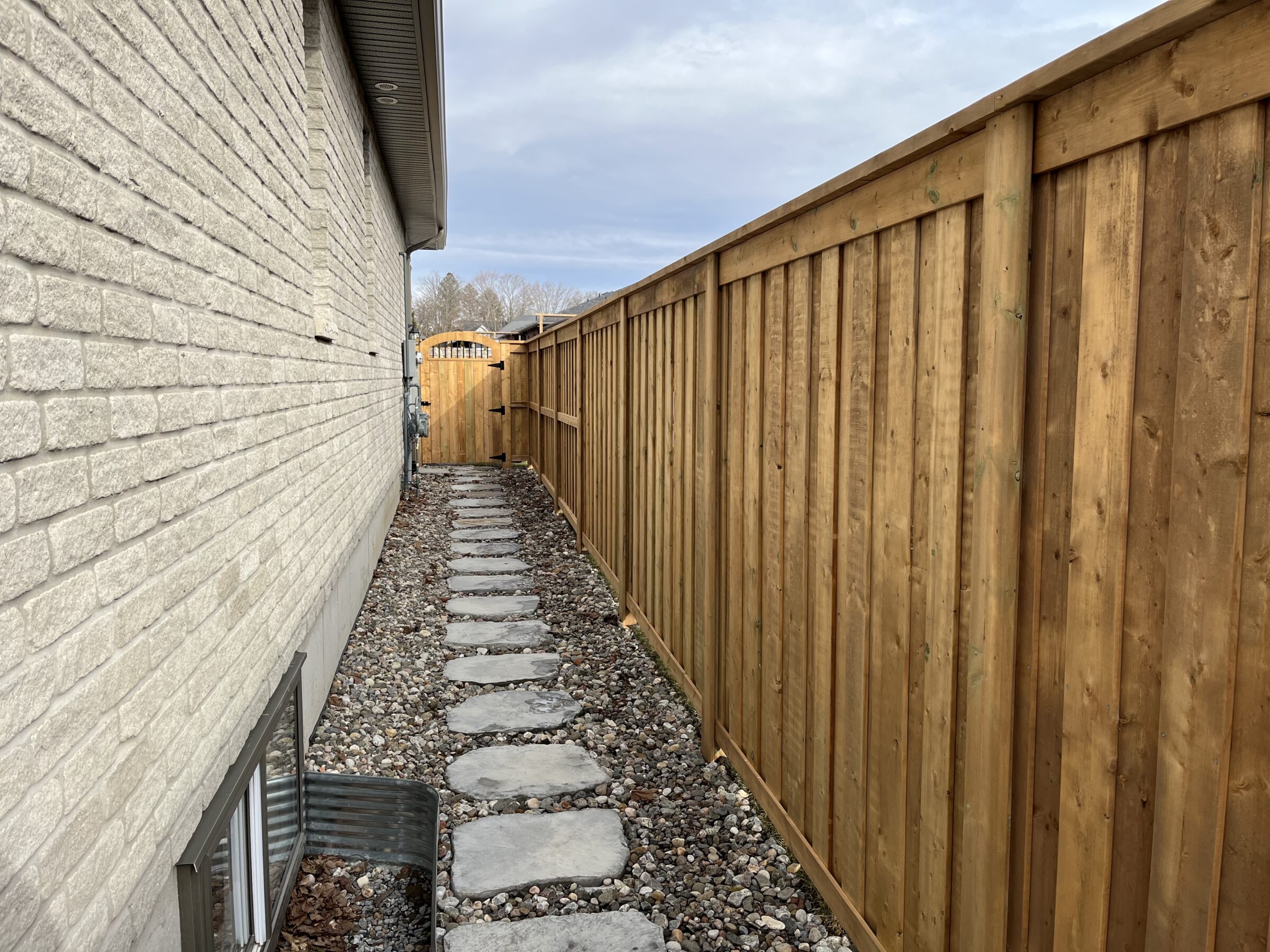 A narrow outdoor pathway with stepping stones, flanked by a brick house wall and wooden fence, leads to a wooden gate.