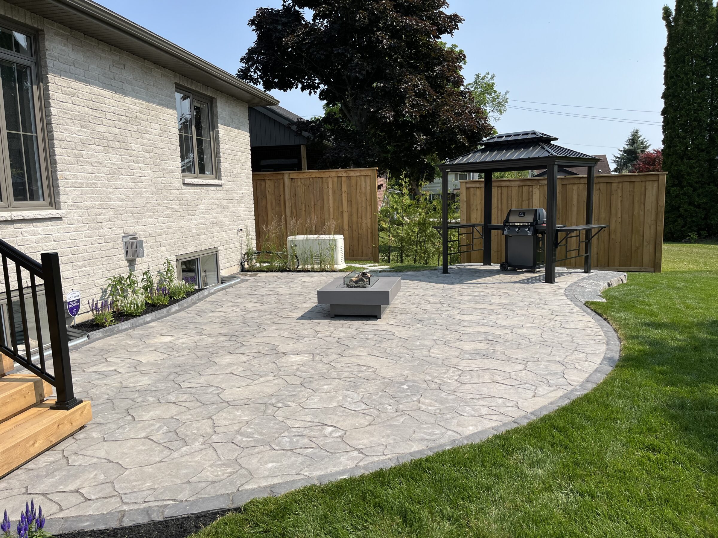 House exterior with stone patio, wooden steps, gazebo covering a grill, manicured lawn, and privacy fence. Plants accent the area.