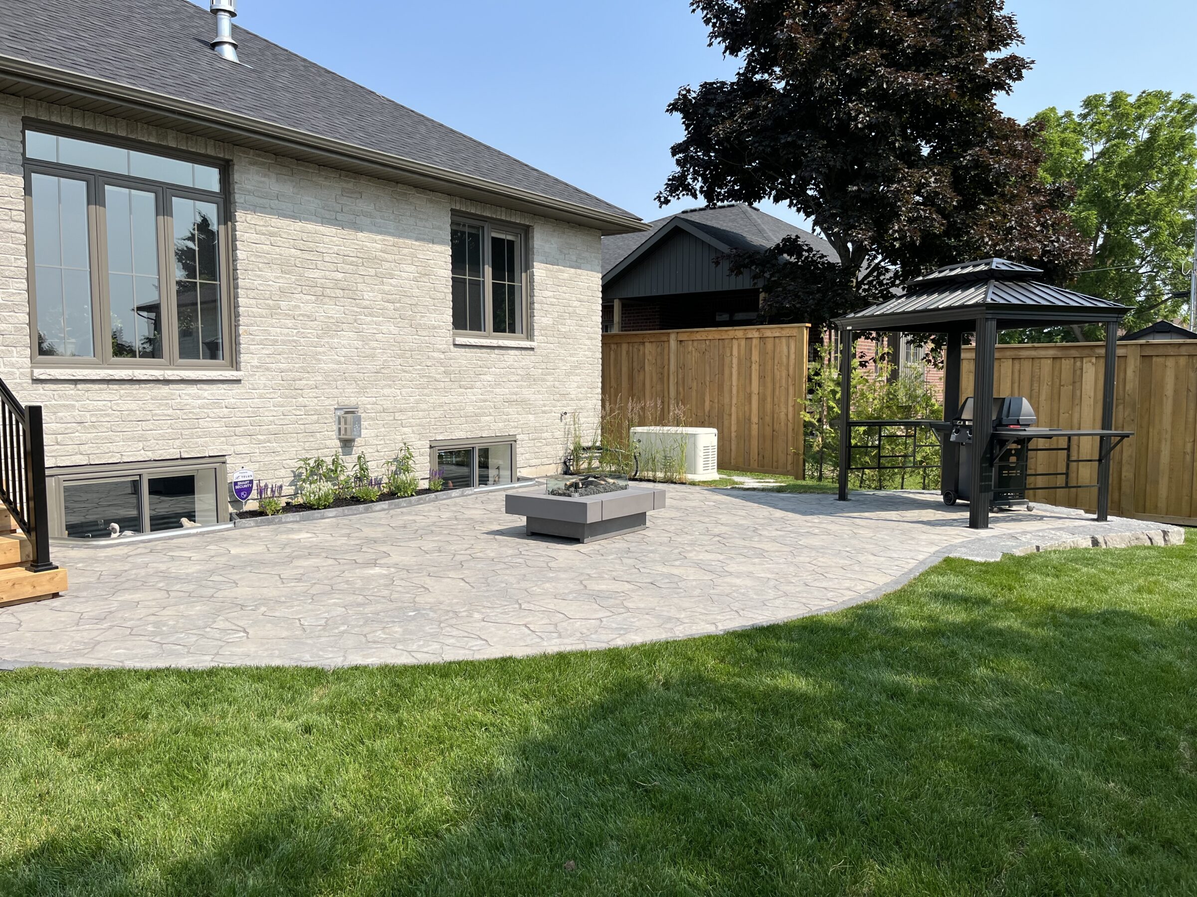 A backyard with a stone patio, gazebo, and grill. Wooden fence, lush grass, and exterior view of a brick house in background.