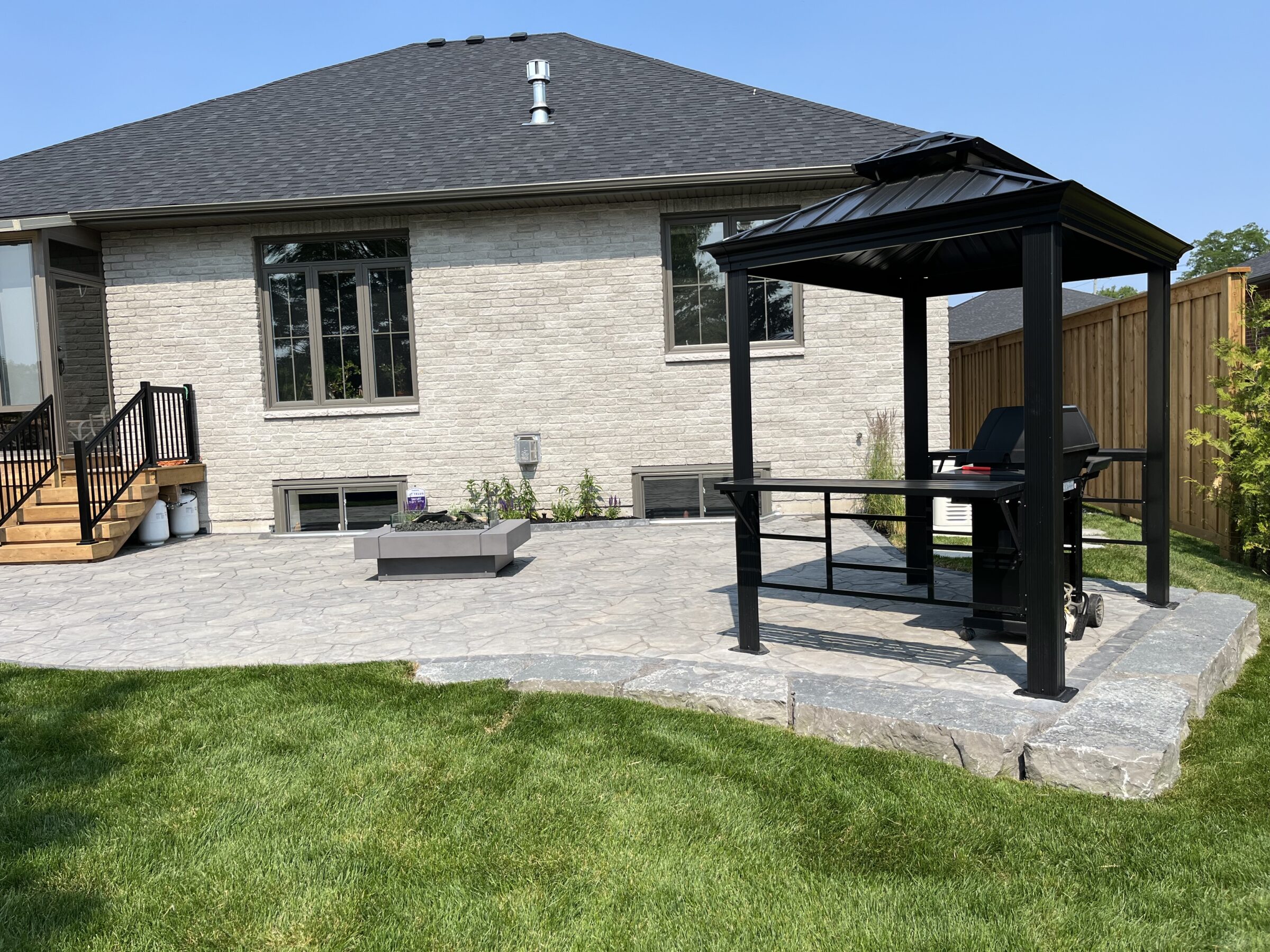 A backyard with a stone patio, gazebo, and grill beside a modern brick house, surrounded by a wooden fence and green lawn.