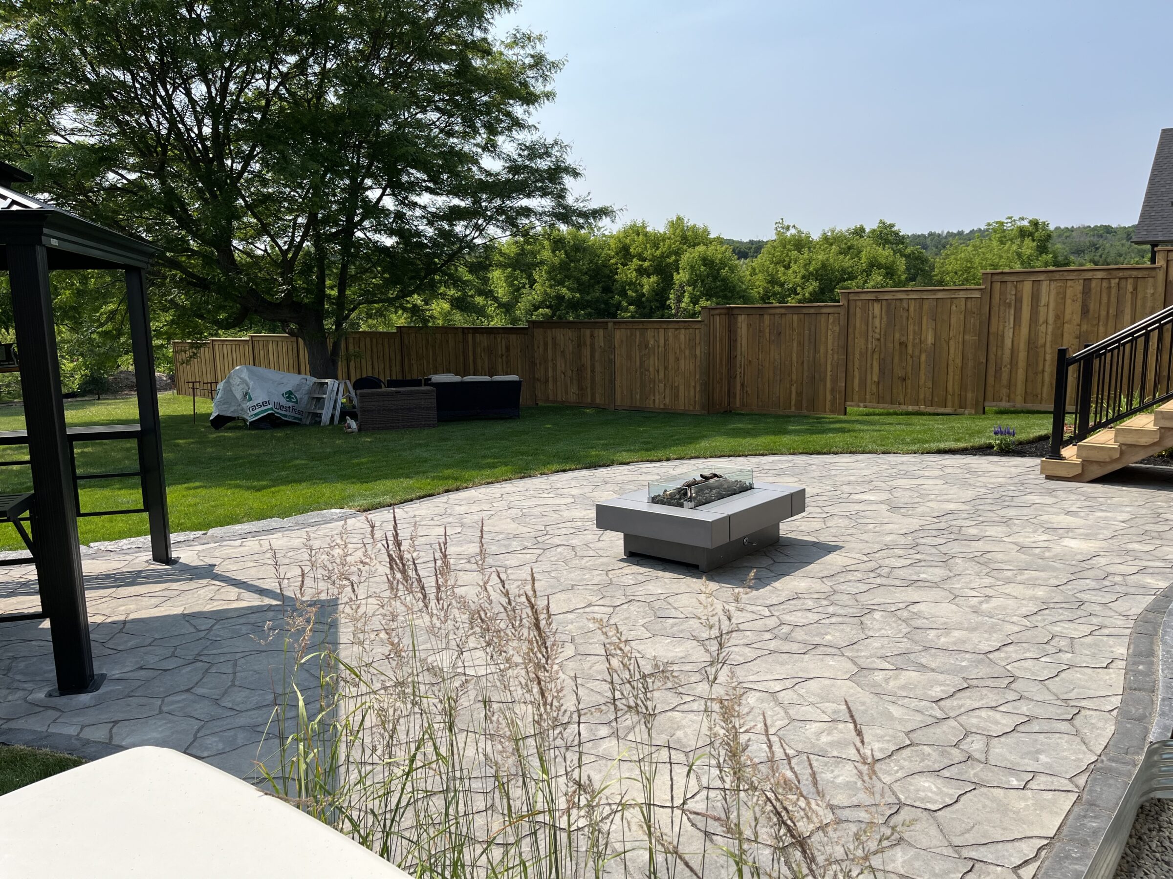 A backyard with a stone patio, modern fire pit, wooden fence, and outdoor furniture under a tree, surrounded by greenery.