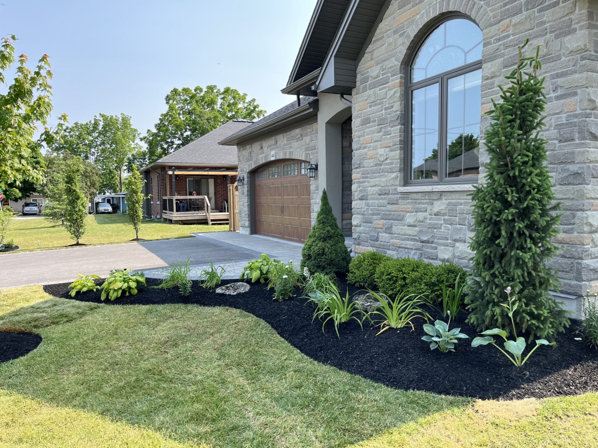 A modern house with stone facade, lush gardens, and driveway. Person is visible on porch of neighboring brick house. Trees in background.