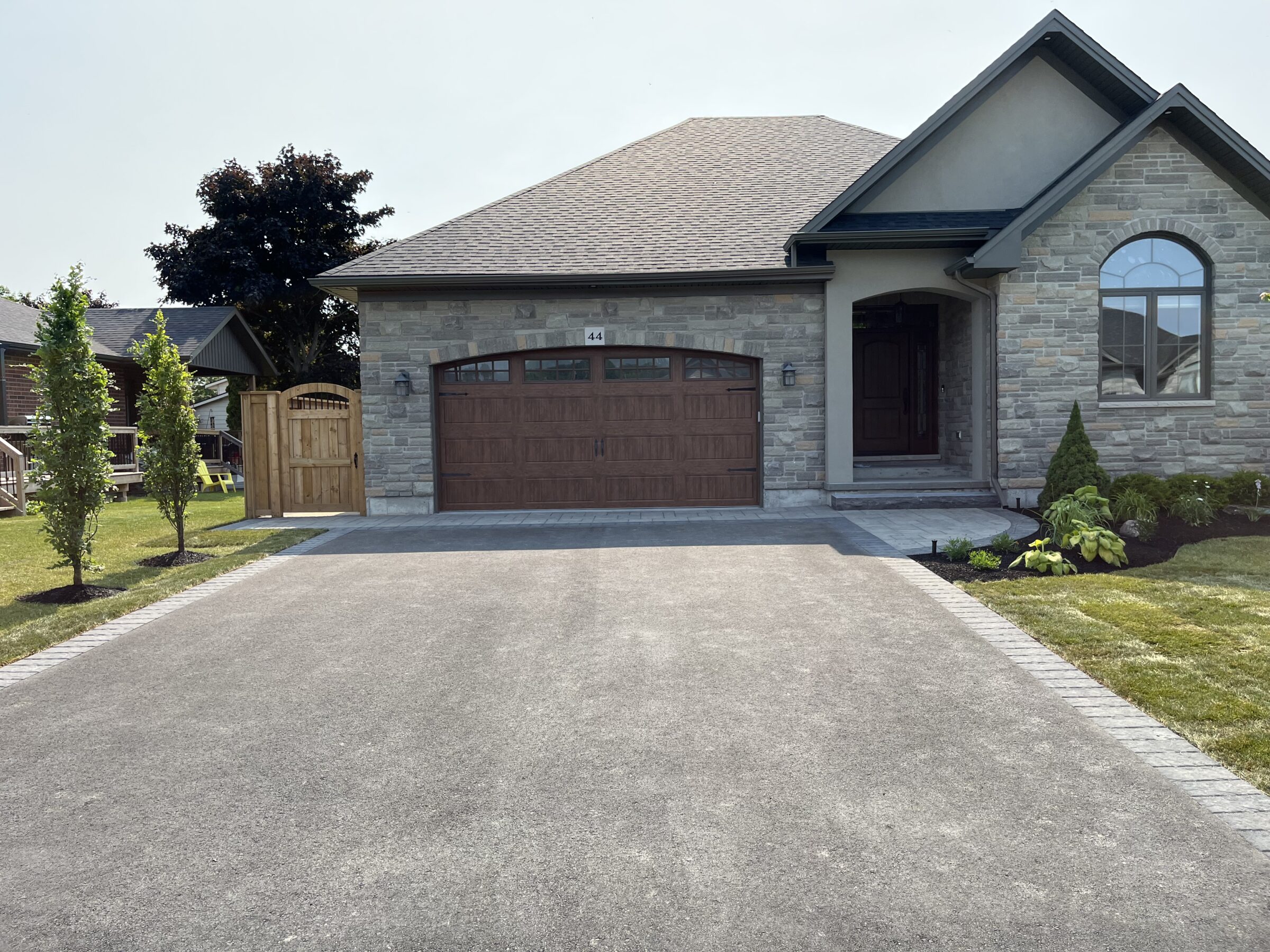 Modern stone house with wooden garage door, manicured lawn, and young trees. A fenced pathway leads to the entrance. Bright, sunny day.