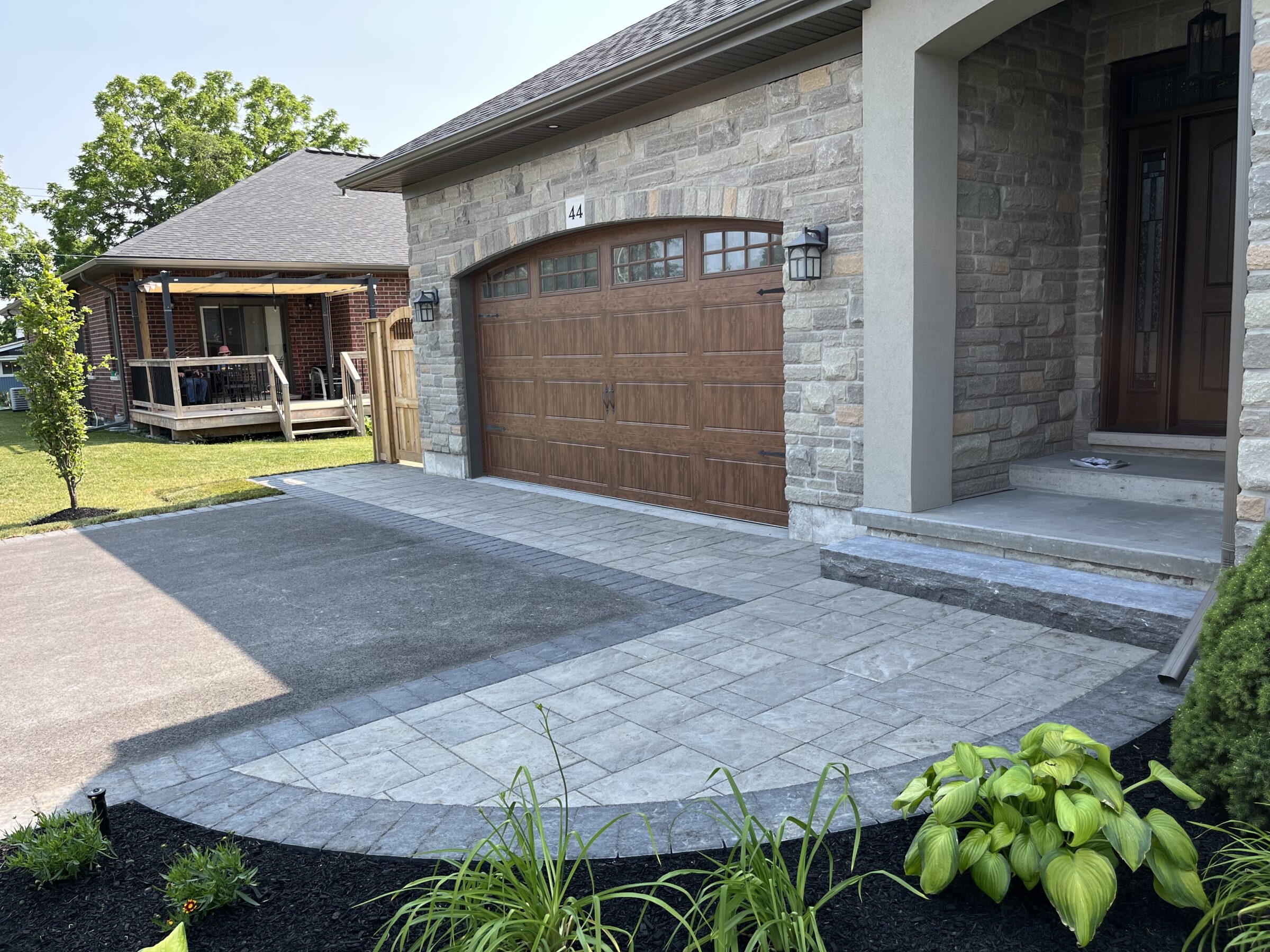 A modern stone house with a wooden garage door, landscaped front yard, and a porch. A person sits on a distant patio.
