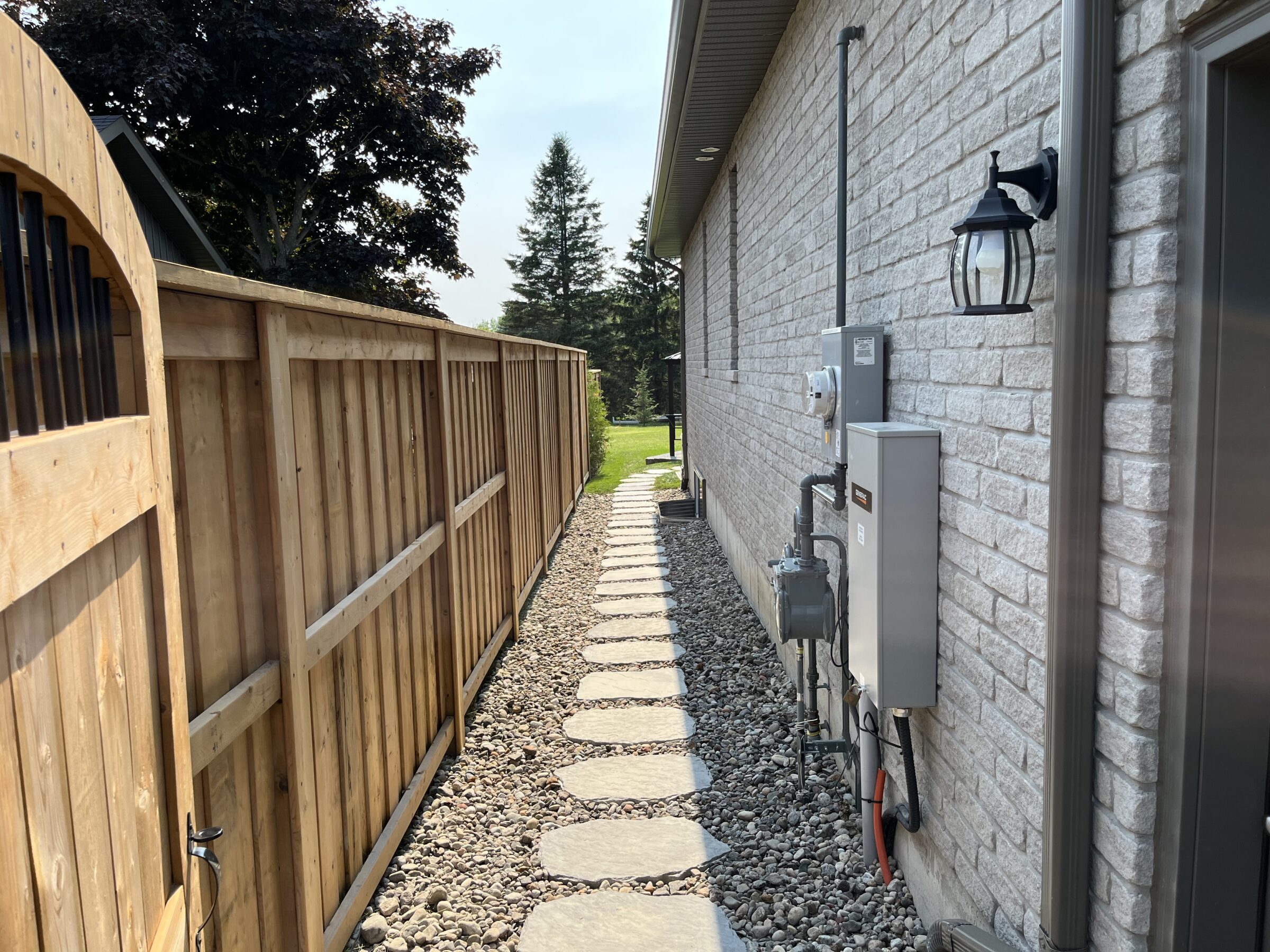 Narrow stone path between a wooden fence and brick house, leading to a grassy yard. Outdoor light and utility meters visible.