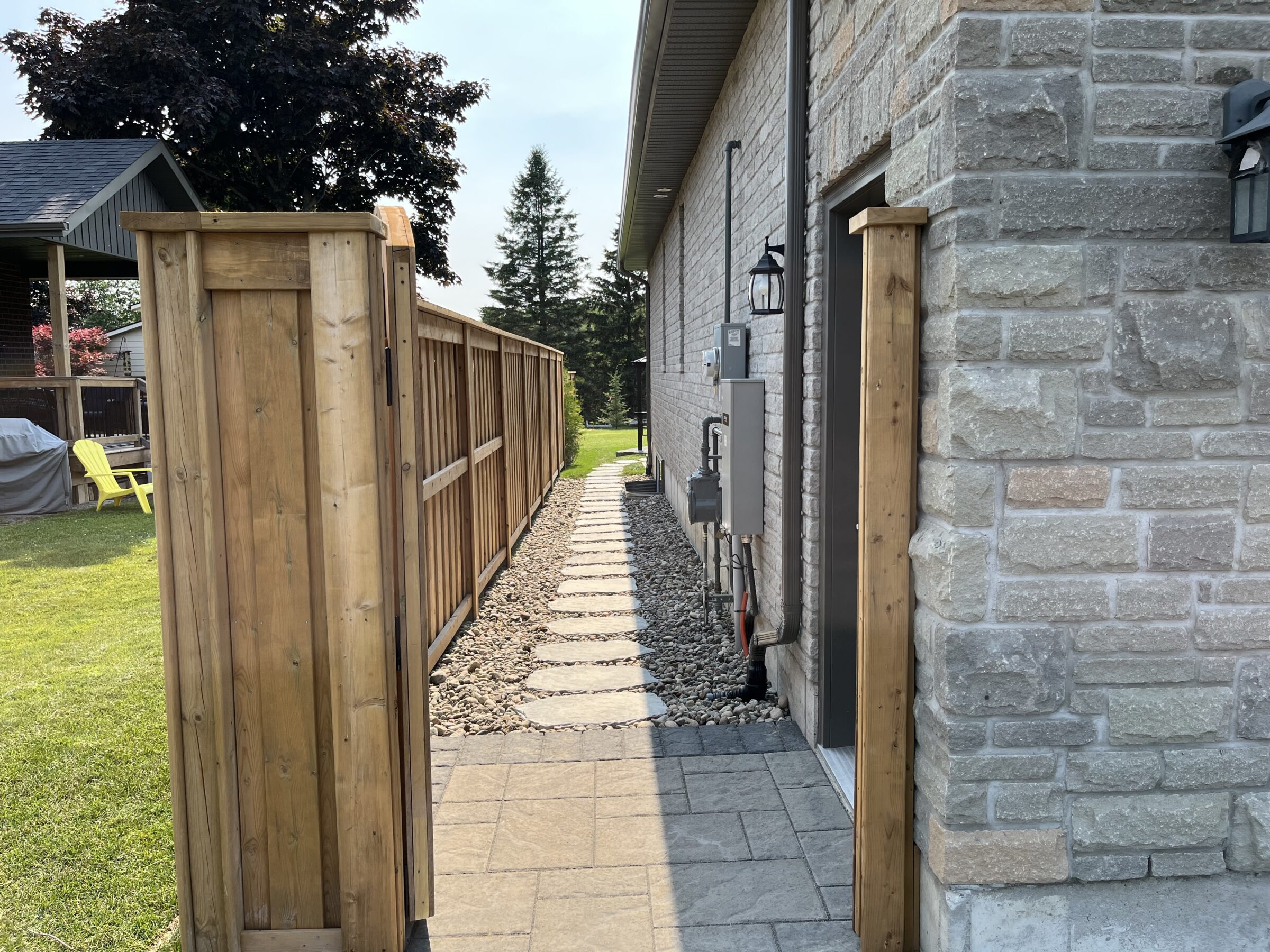 A wooden fence and stone walkway run alongside a brick house, leading to a green backyard with trees and outdoor seating.