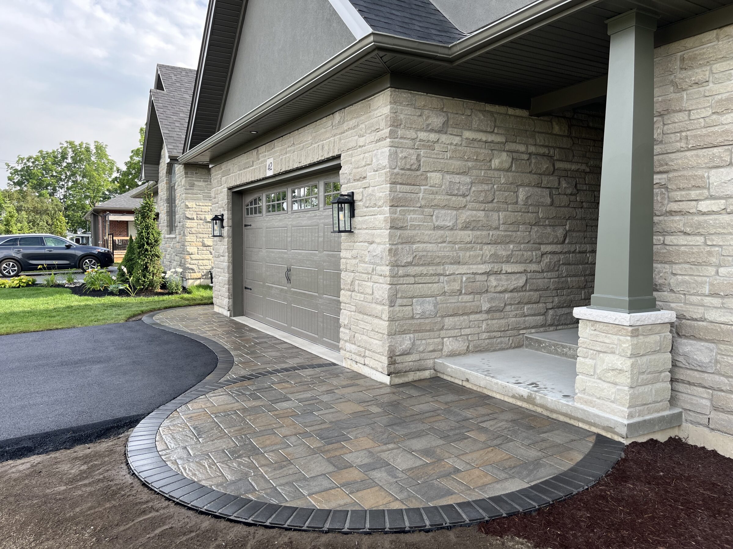 Modern suburban house with stone exterior, paved driveway, and well-maintained lawn. A black car is parked nearby. Overcast sky in the background.