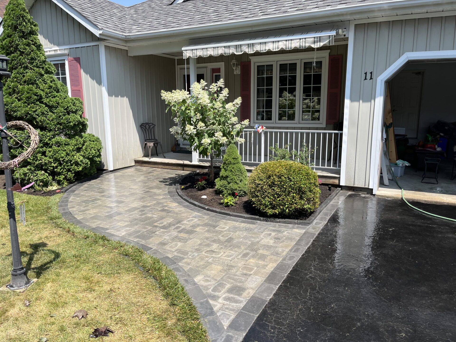 A quaint house with a manicured garden, tiled walkway, and an open garage. A lamppost stands on the green lawn.