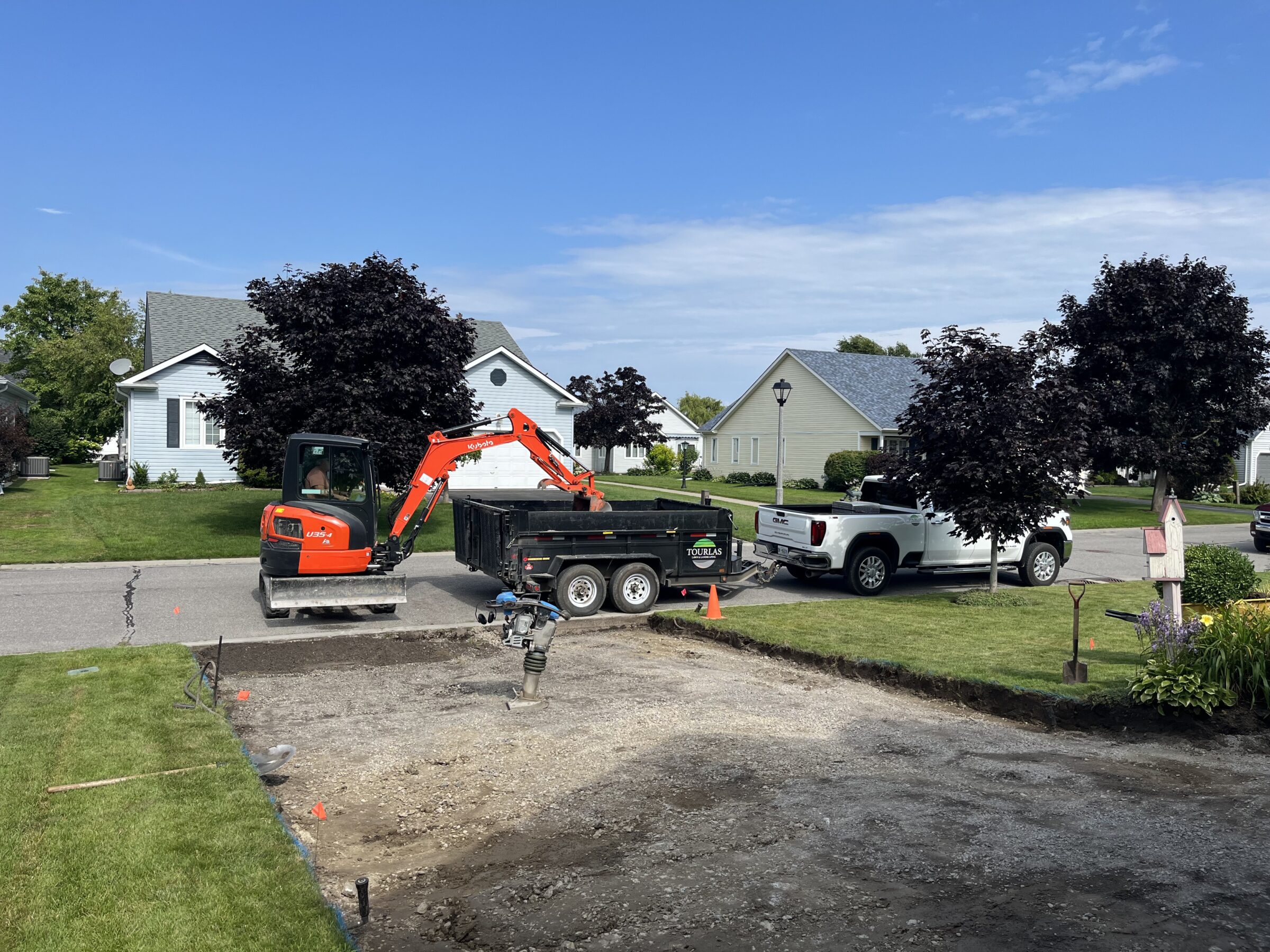 A person operates an orange excavator in a suburban neighborhood, preparing a driveway beside a white truck. Houses and trees surround the scene.
