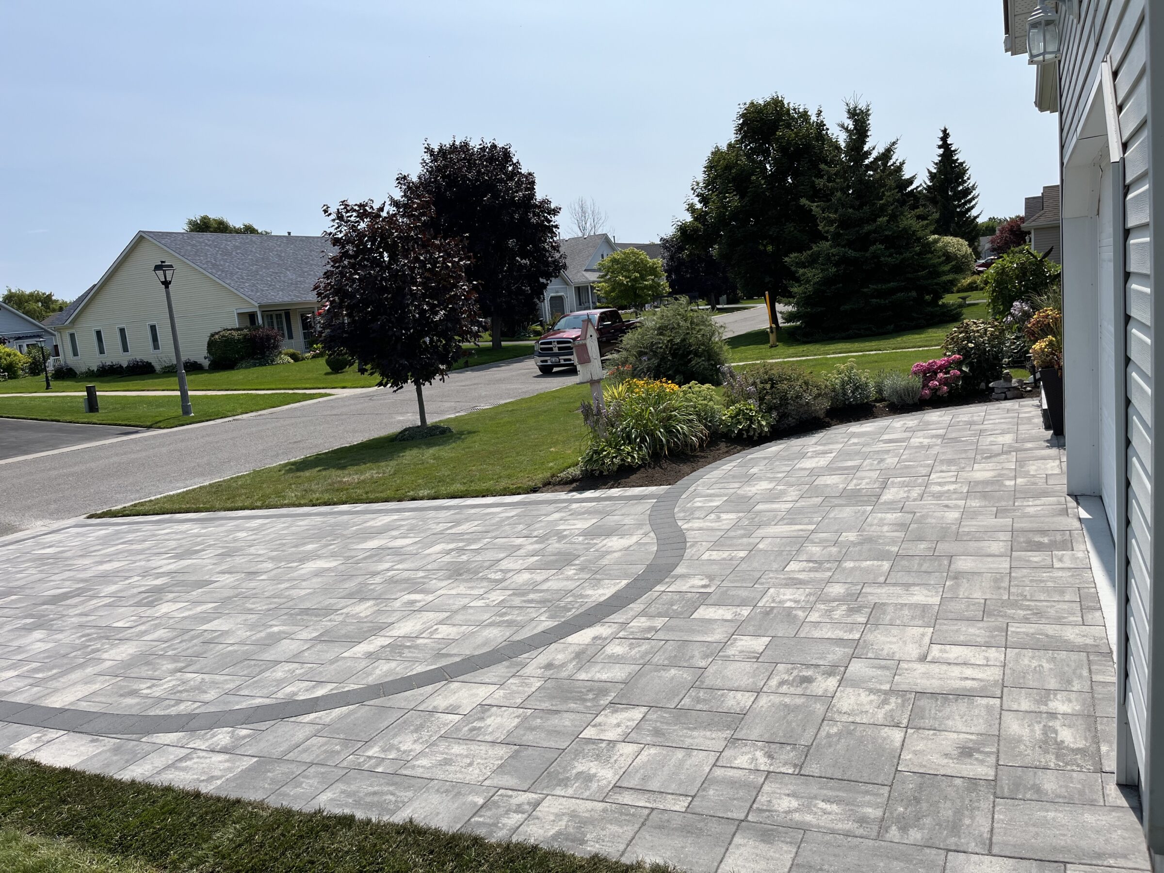 A suburban neighborhood with a newly paved driveway, well-maintained lawns, and lush gardens, featuring parked cars and single-story houses under a clear sky.