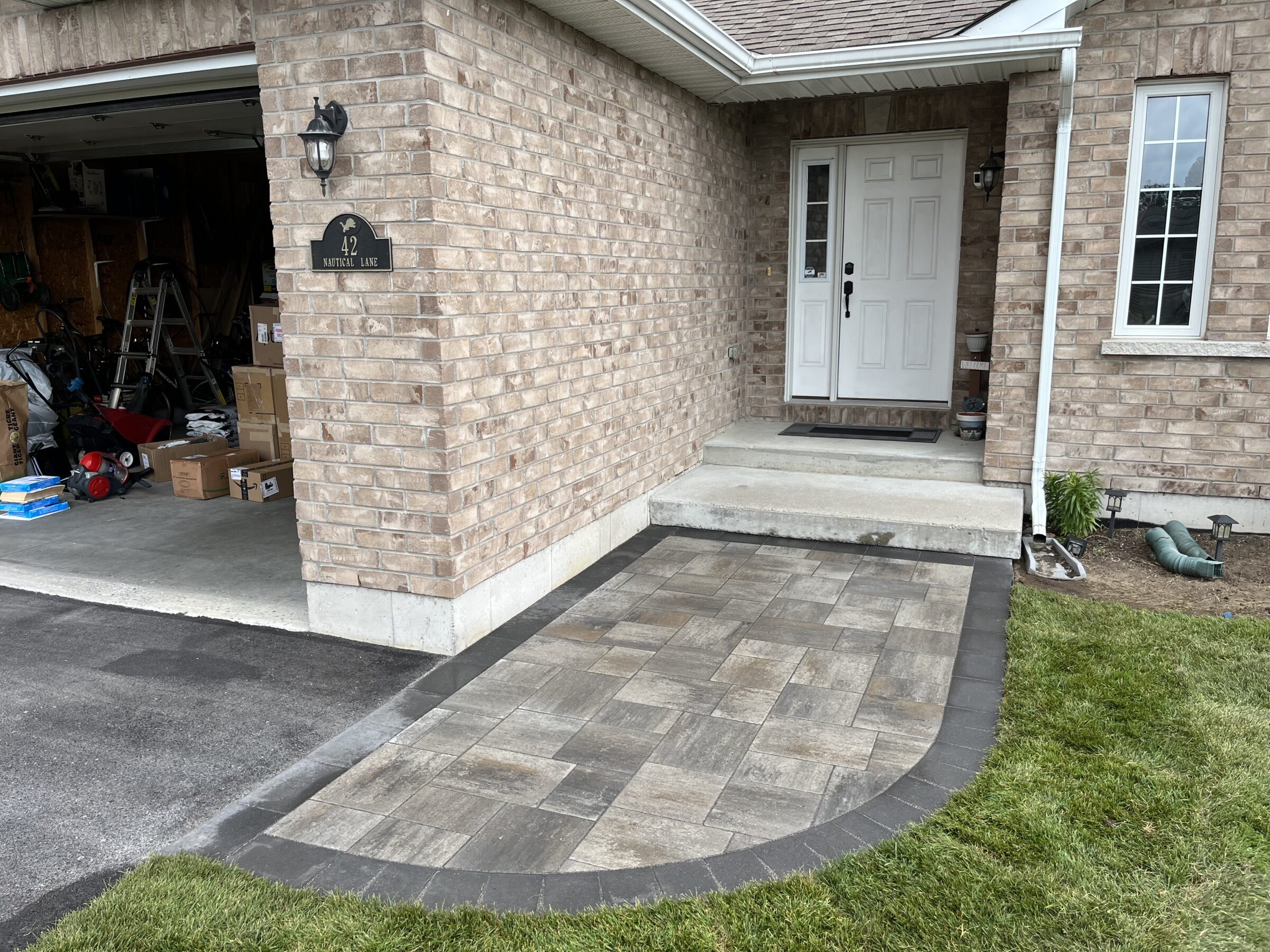 A brick house with a white door, open garage, visible boxes, lawn, and a new stone walkway. No people or landmarks present.