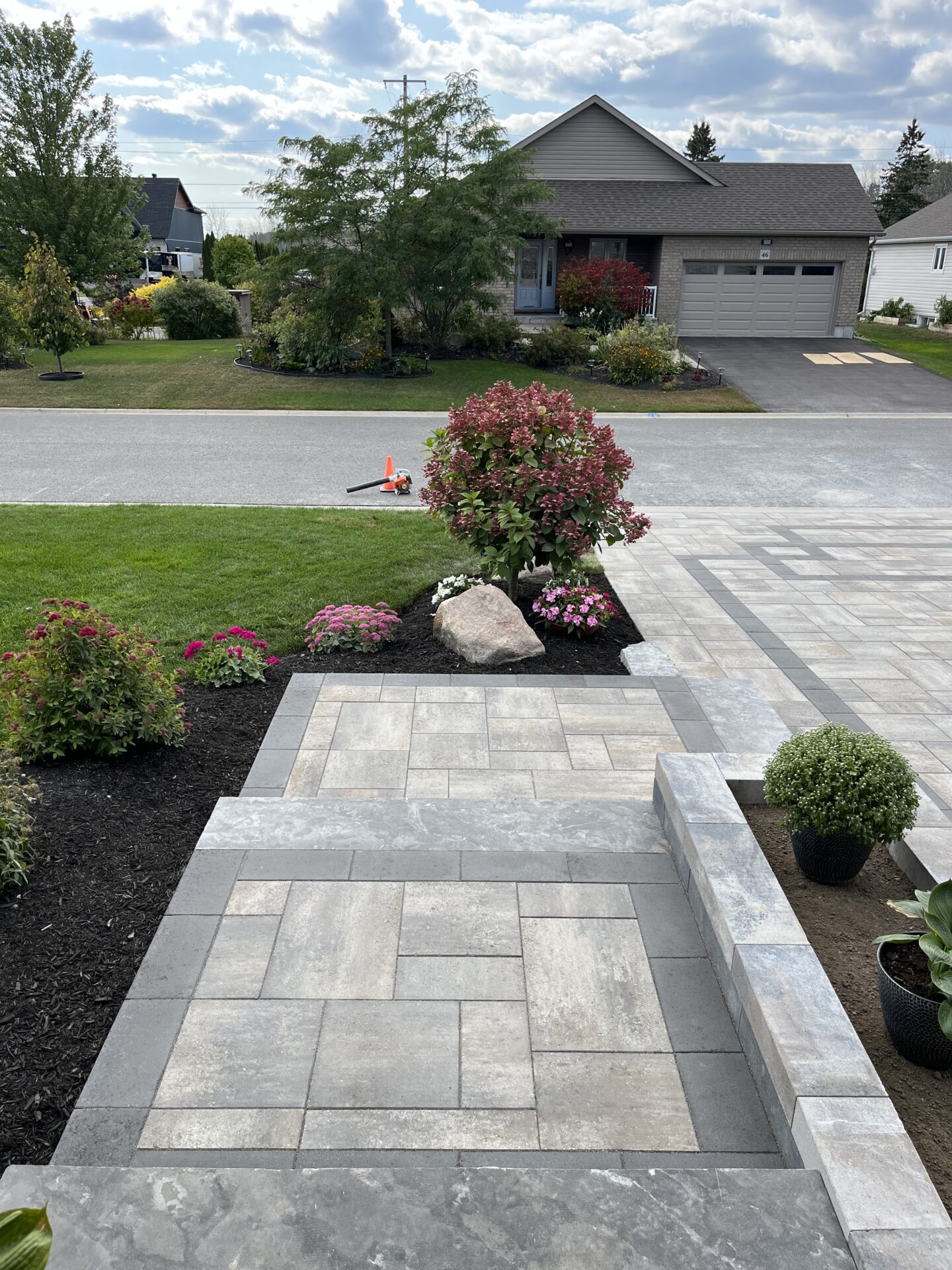 A paved walkway leads to a suburban house with a well-maintained garden, featuring colorful flowers and a few scattered tools.