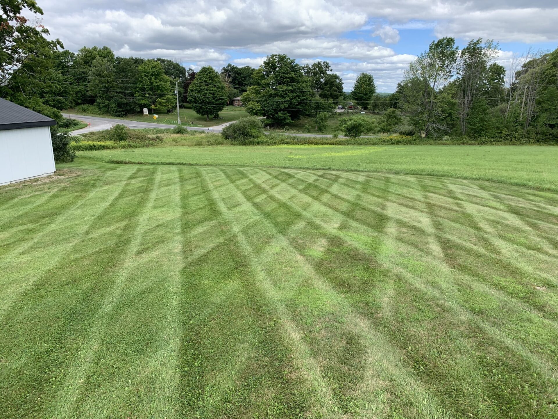 A freshly mowed lawn with a crisscross pattern, surrounded by green trees and a road in a rural landscape under a partly cloudy sky.