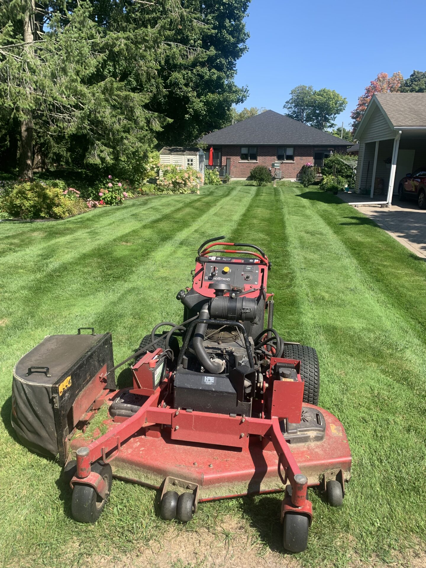 A red lawn mower sits on a freshly cut, lush green lawn, with a red brick house and trees in the background.