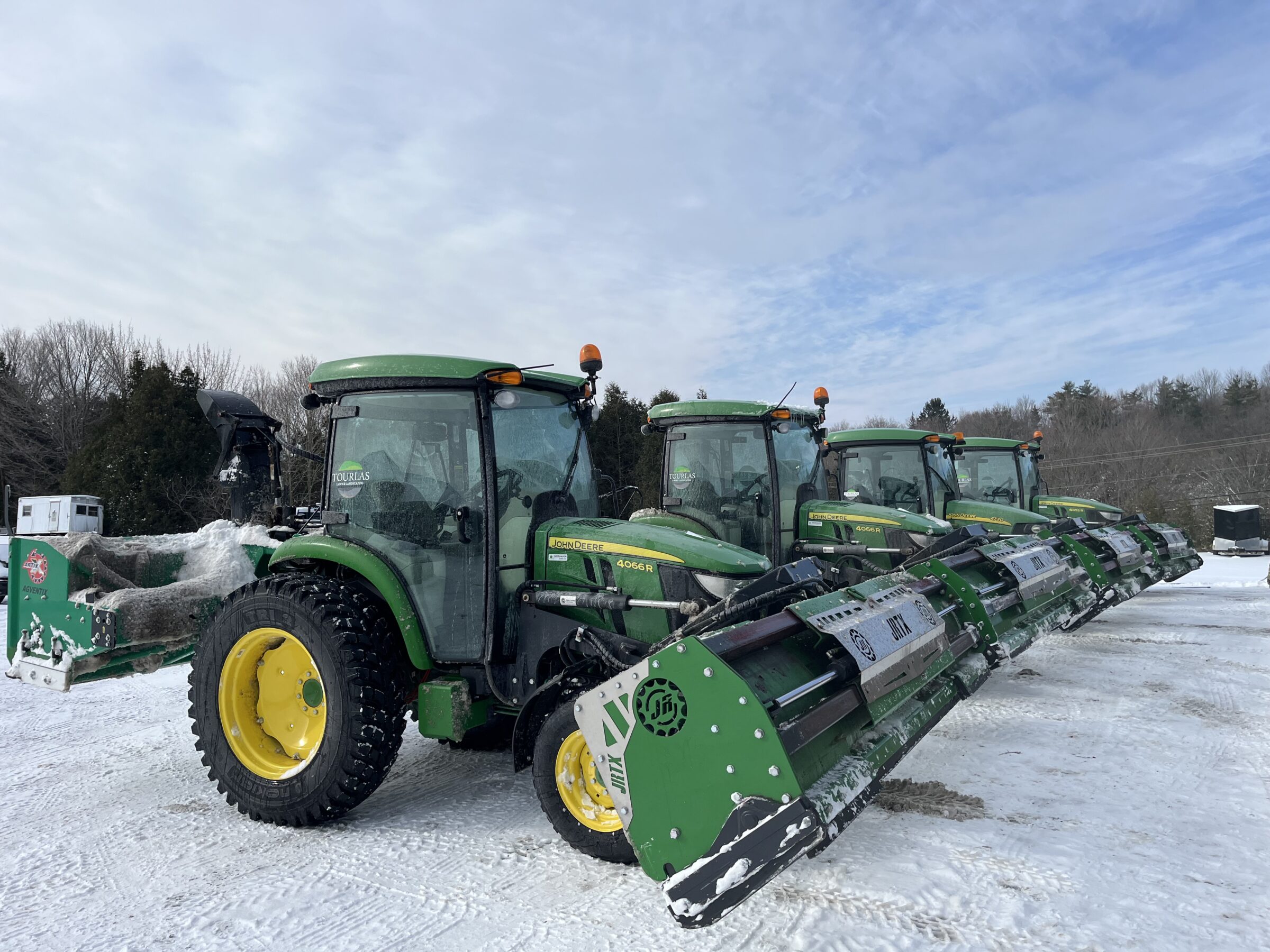 A row of green tractors with plows line up on snowy ground, surrounded by trees under a cloudy sky.
