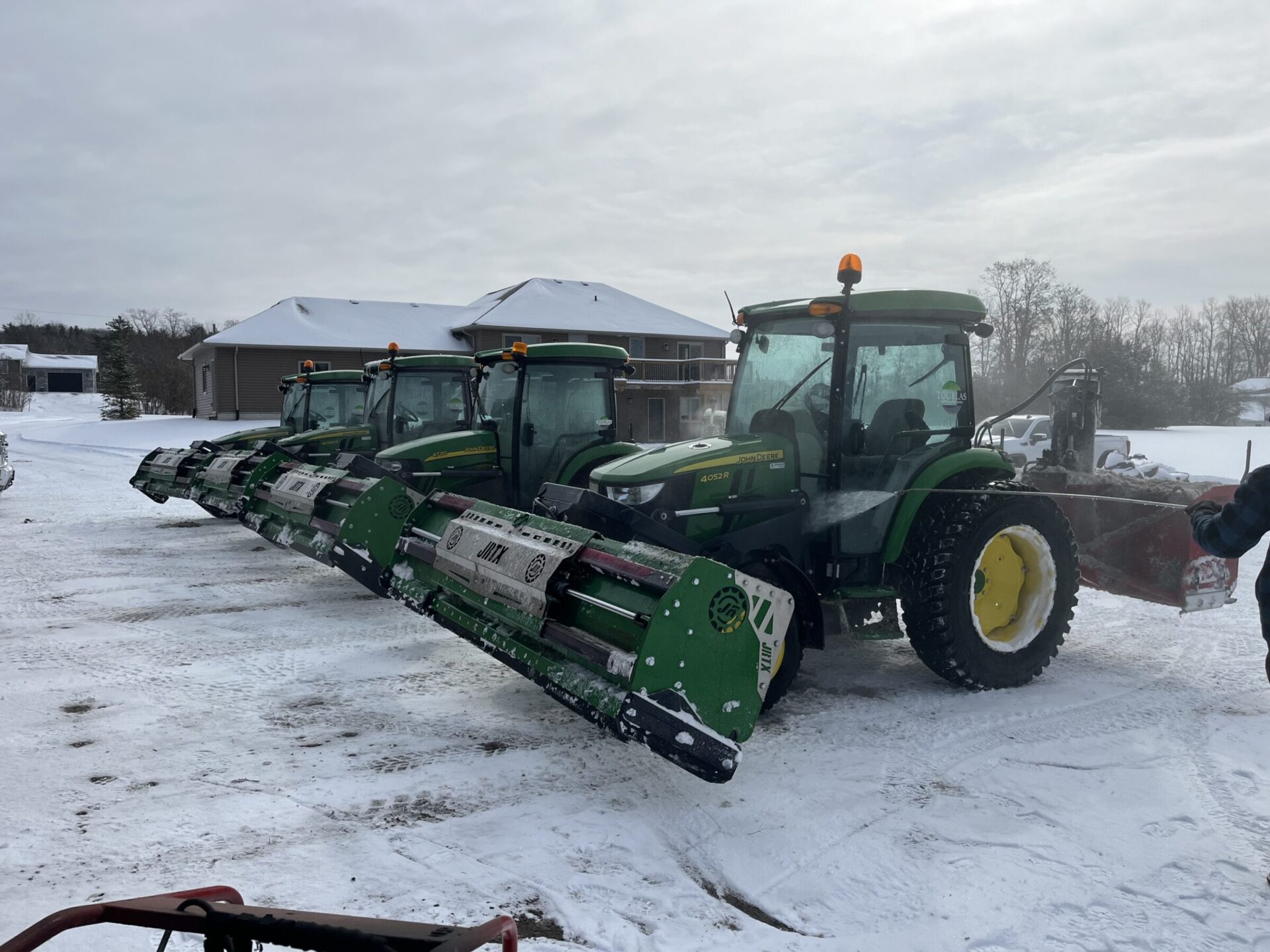 Four tractors with green snow plows parked on snowy ground beside a building, while a person inspects them under a cloudy sky.