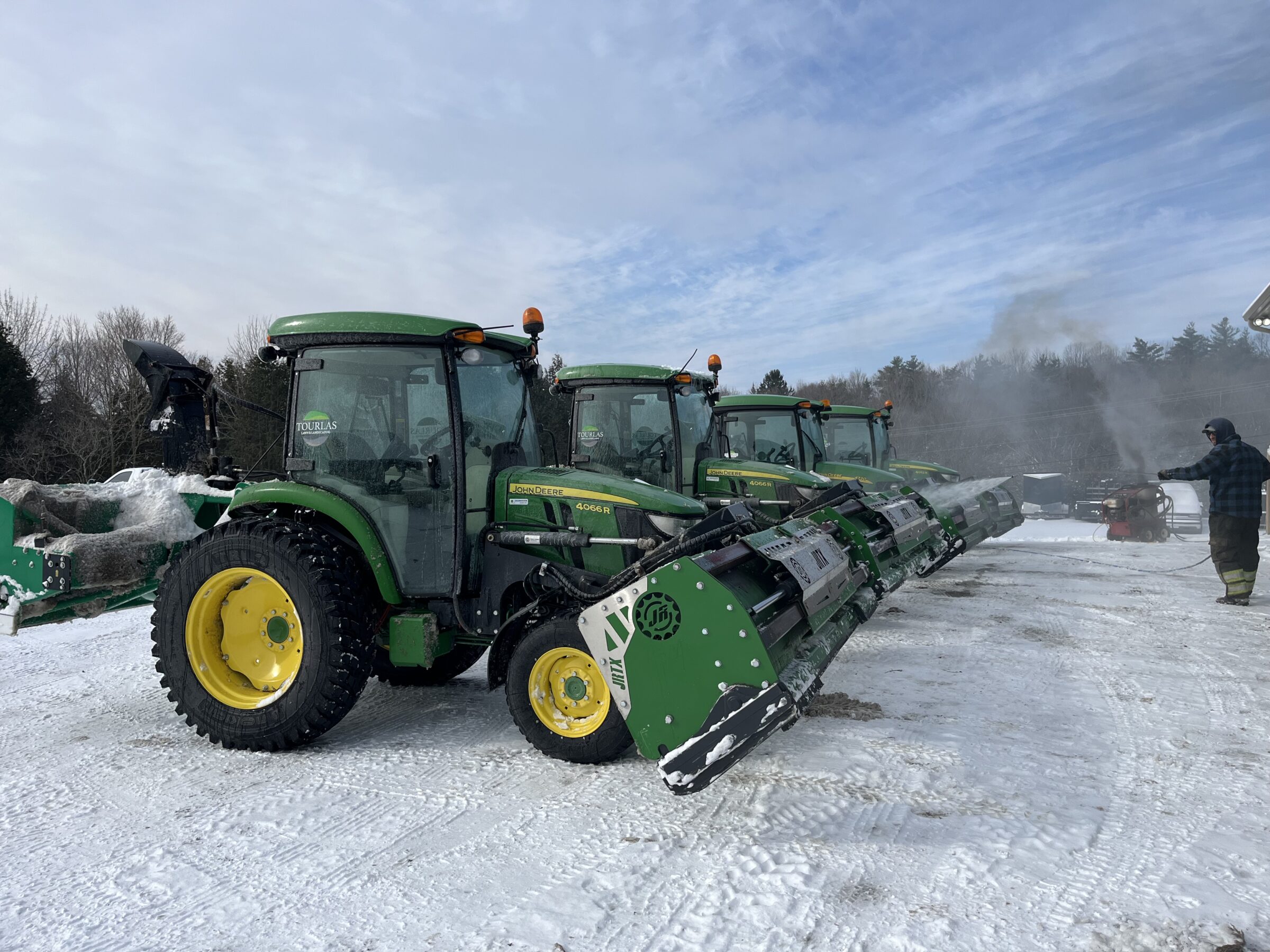 Several tractors are lined up on a snowy surface. A person wearing winter clothing operates machinery, with trees visible in the background.
