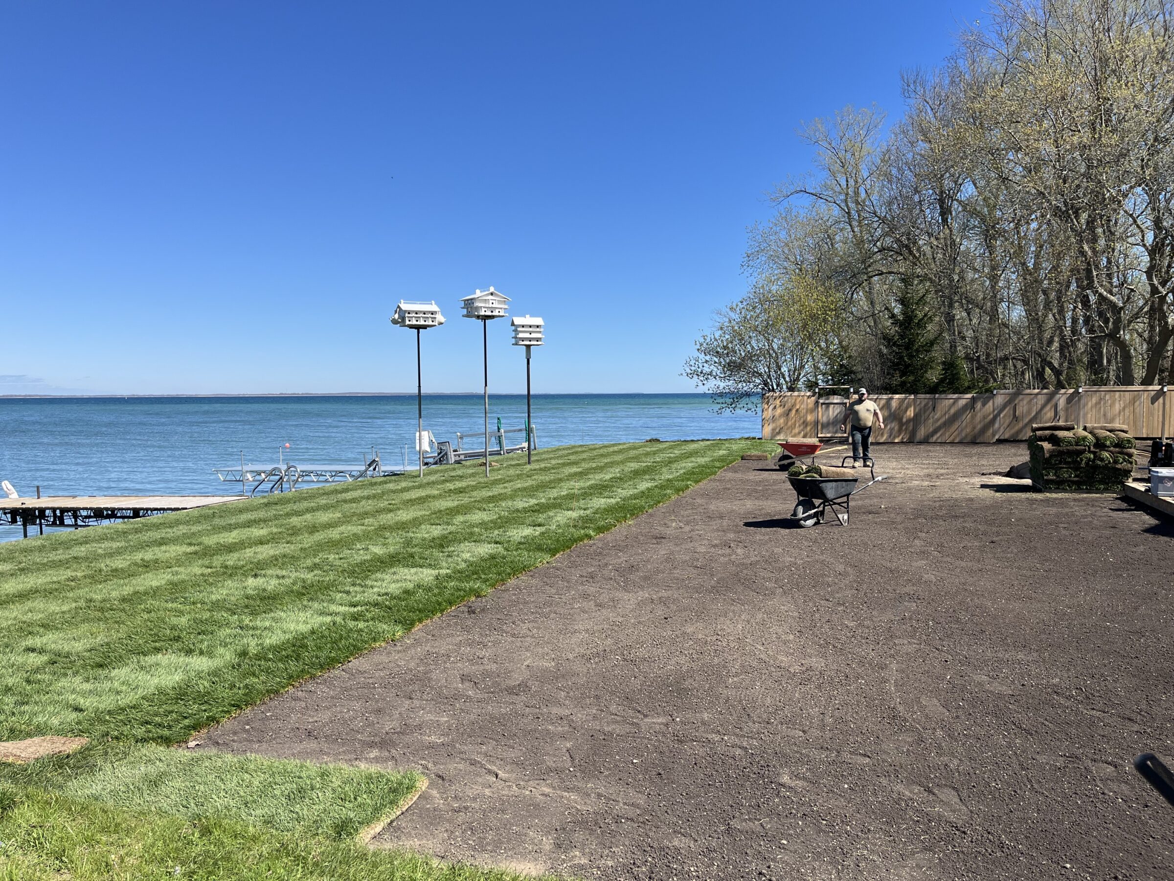 A person with a wheelbarrow works near a lake. Birdhouses, a dock, grass, and trees are visible under a clear blue sky.