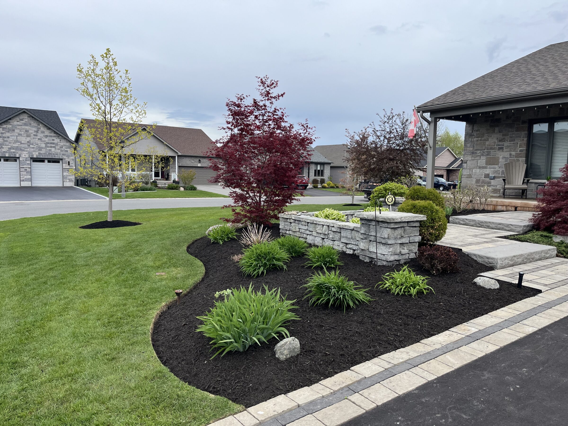 A suburban neighborhood scene featuring a well-manicured garden, stone pathway, and Canadian flag. Modern houses and neatly trimmed lawns surround the area.
