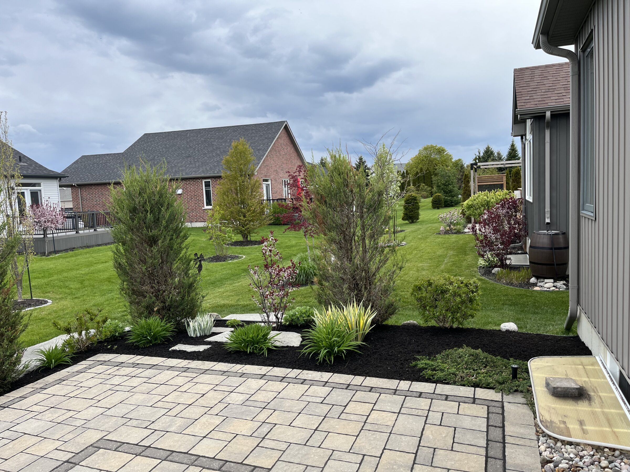 Backyard with brick house, manicured lawn, paver patio, and neatly planted shrubs. Overcast sky creates a serene suburban atmosphere.
