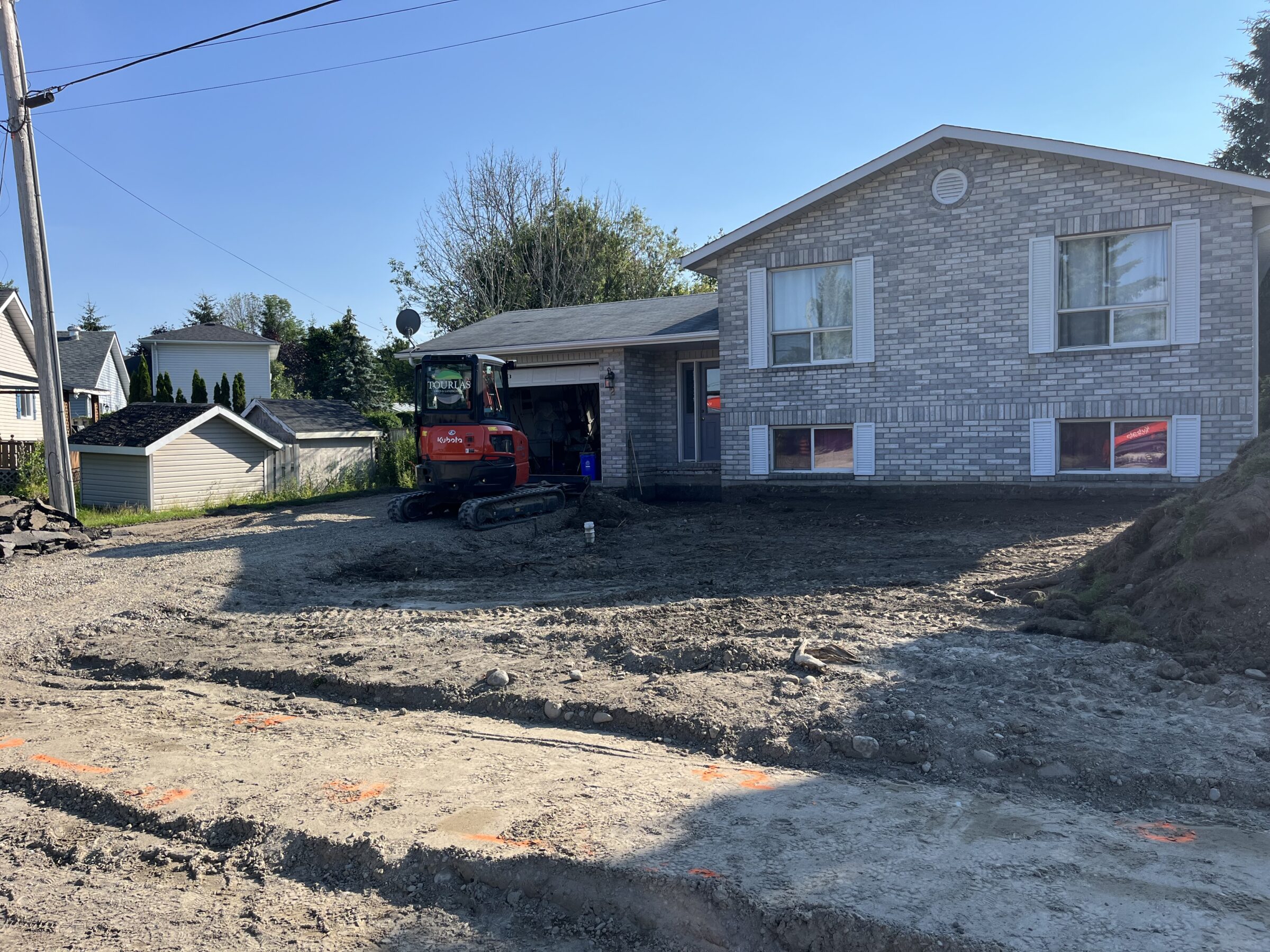 A brick house with construction equipment is undergoing landscaping. Nearby are smaller structures and some trees under a clear blue sky.