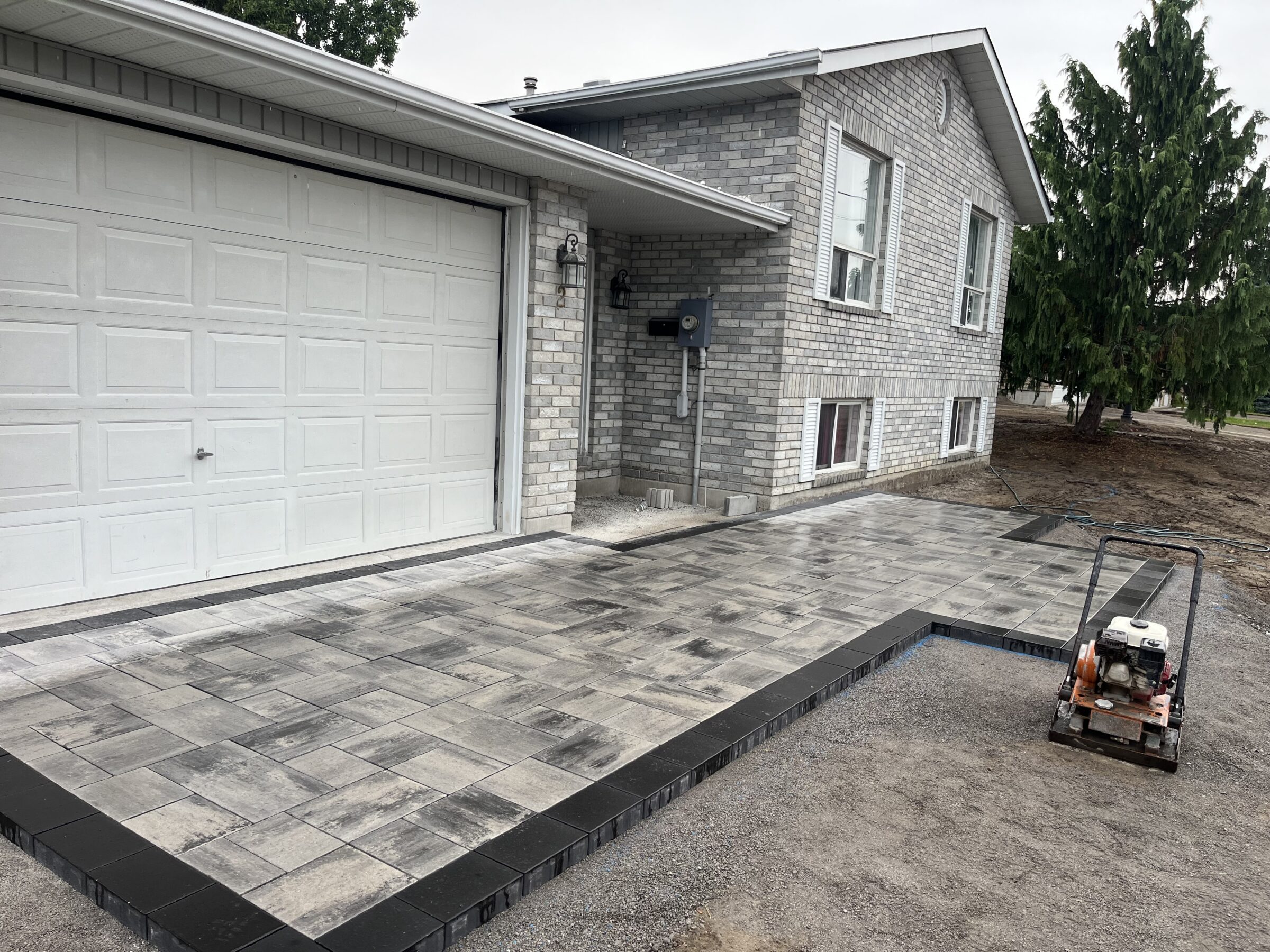 A newly paved driveway with interlocking stones leads to a gray brick house and attached garage, adjacent to a lush tree.