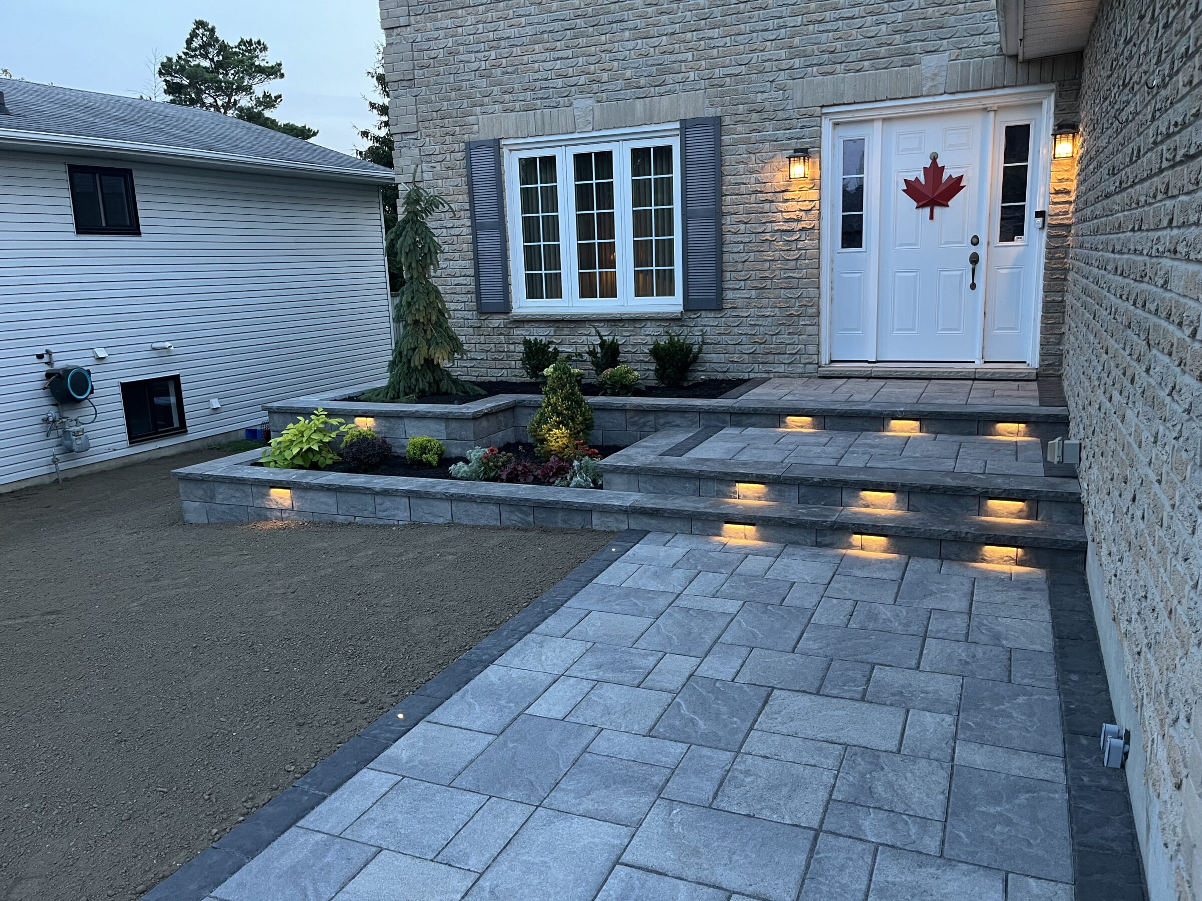 A modern house entrance with steps and illuminated pathway, features a Canadian flag decoration on the front door. Neat landscaping enhances curb appeal.