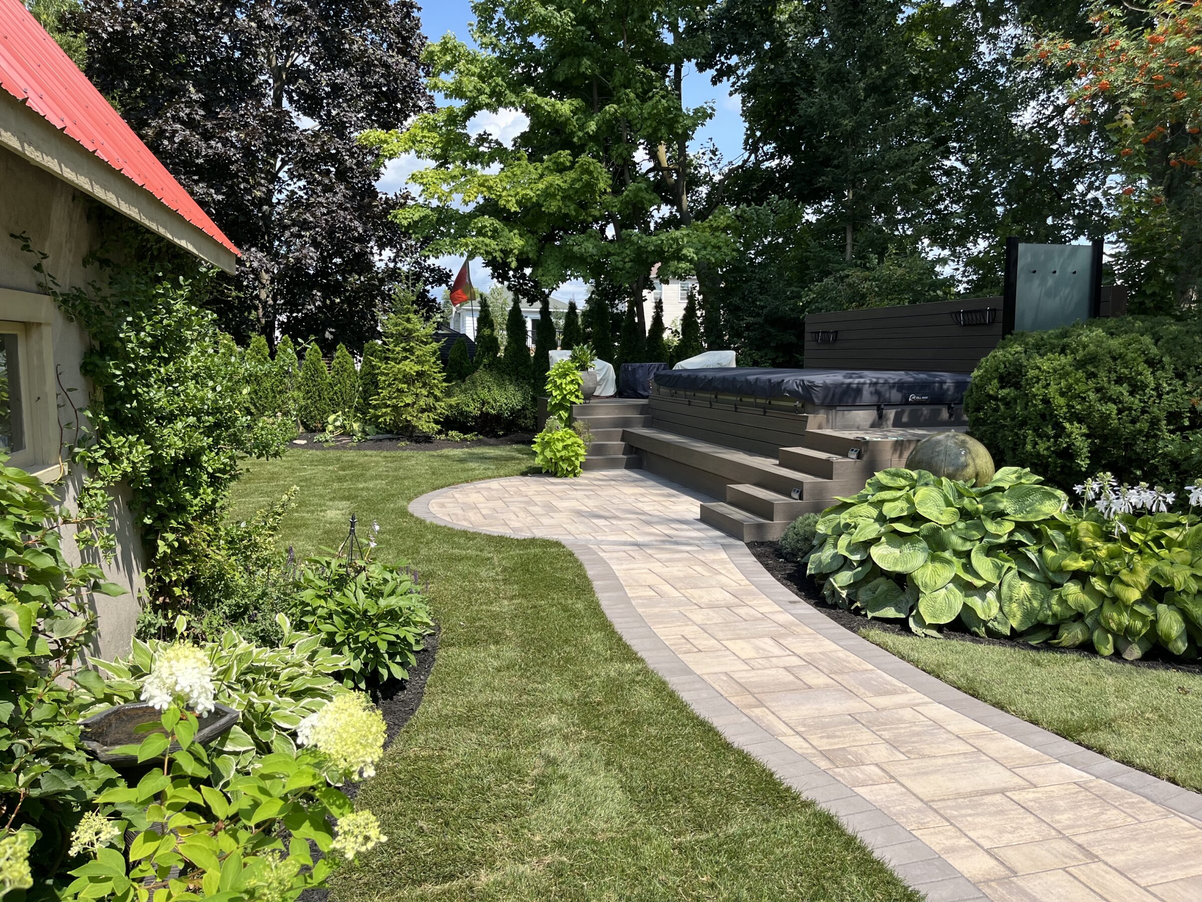 A beautifully landscaped garden with a winding stone pathway, lush greenery, a hot tub, and a red-roofed structure in sunlight.